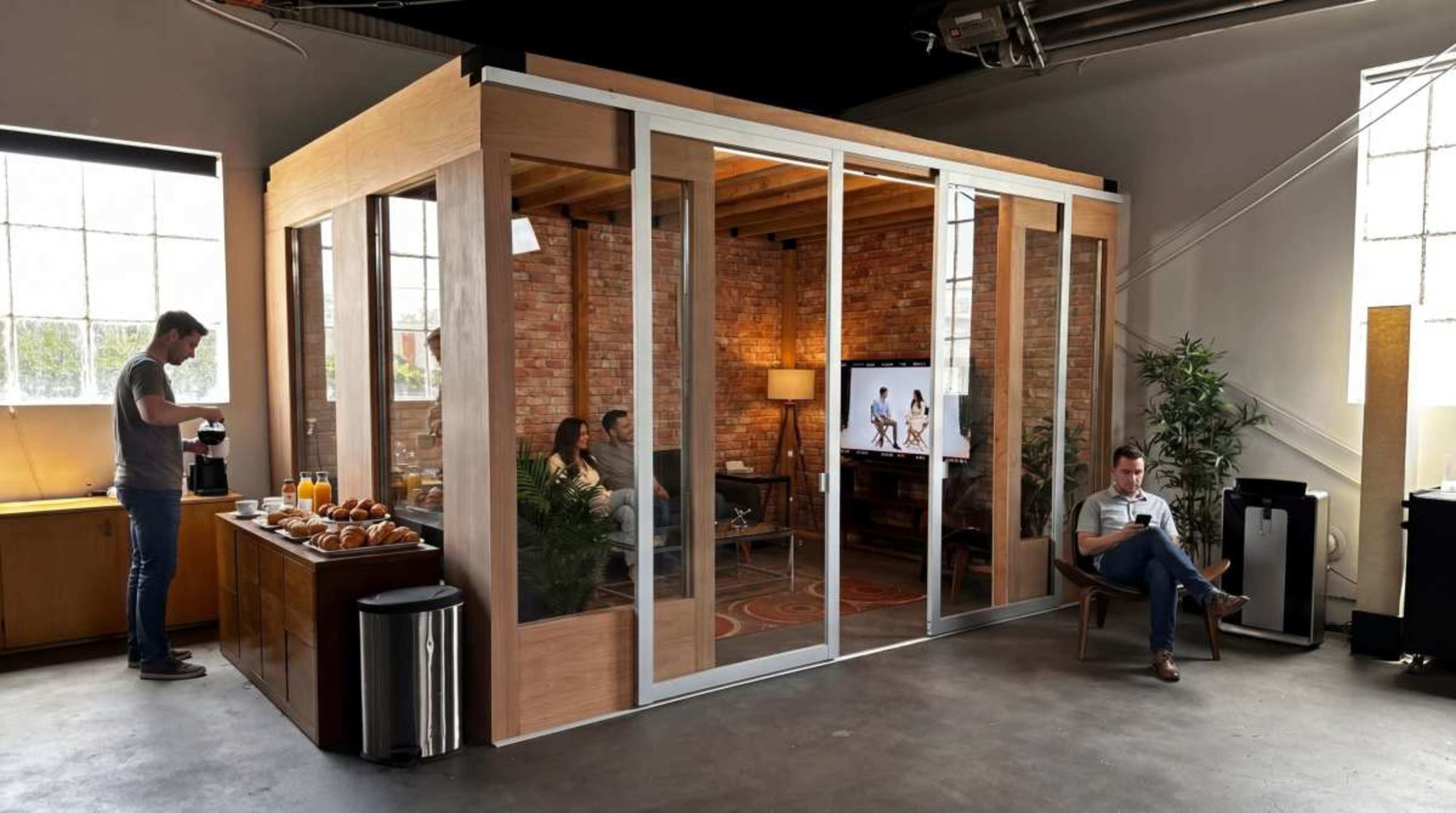 The image shows a modern office space with a glass-walled meeting room, a coffee station with pastries, and people seated inside while another individual prepares coffee nearby.