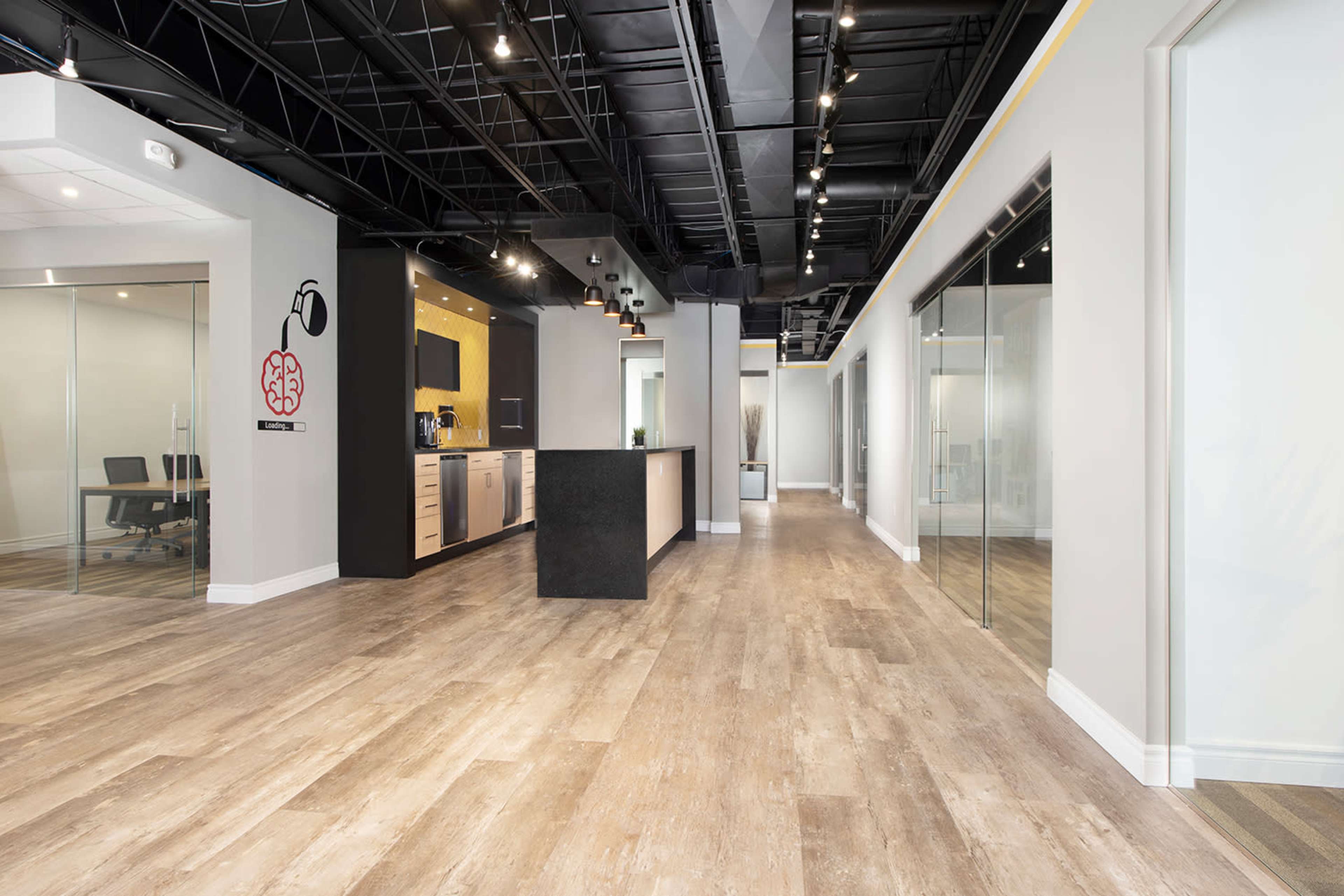 A modern office interior with a central dark island counter, glass-walled meeting rooms, and wooden flooring.