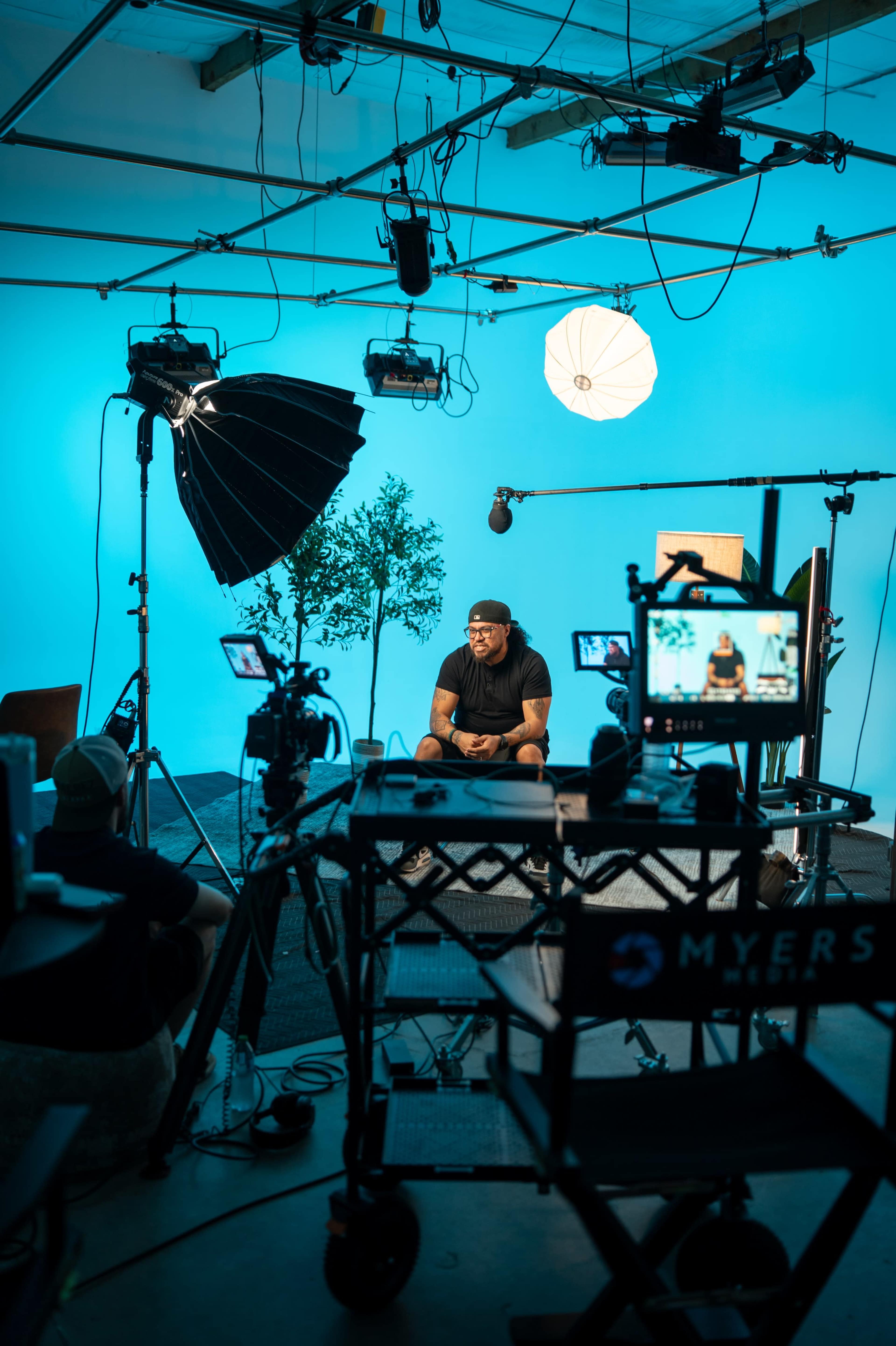 A person sits in front of cameras and lighting equipment in a studio with a blue backdrop.