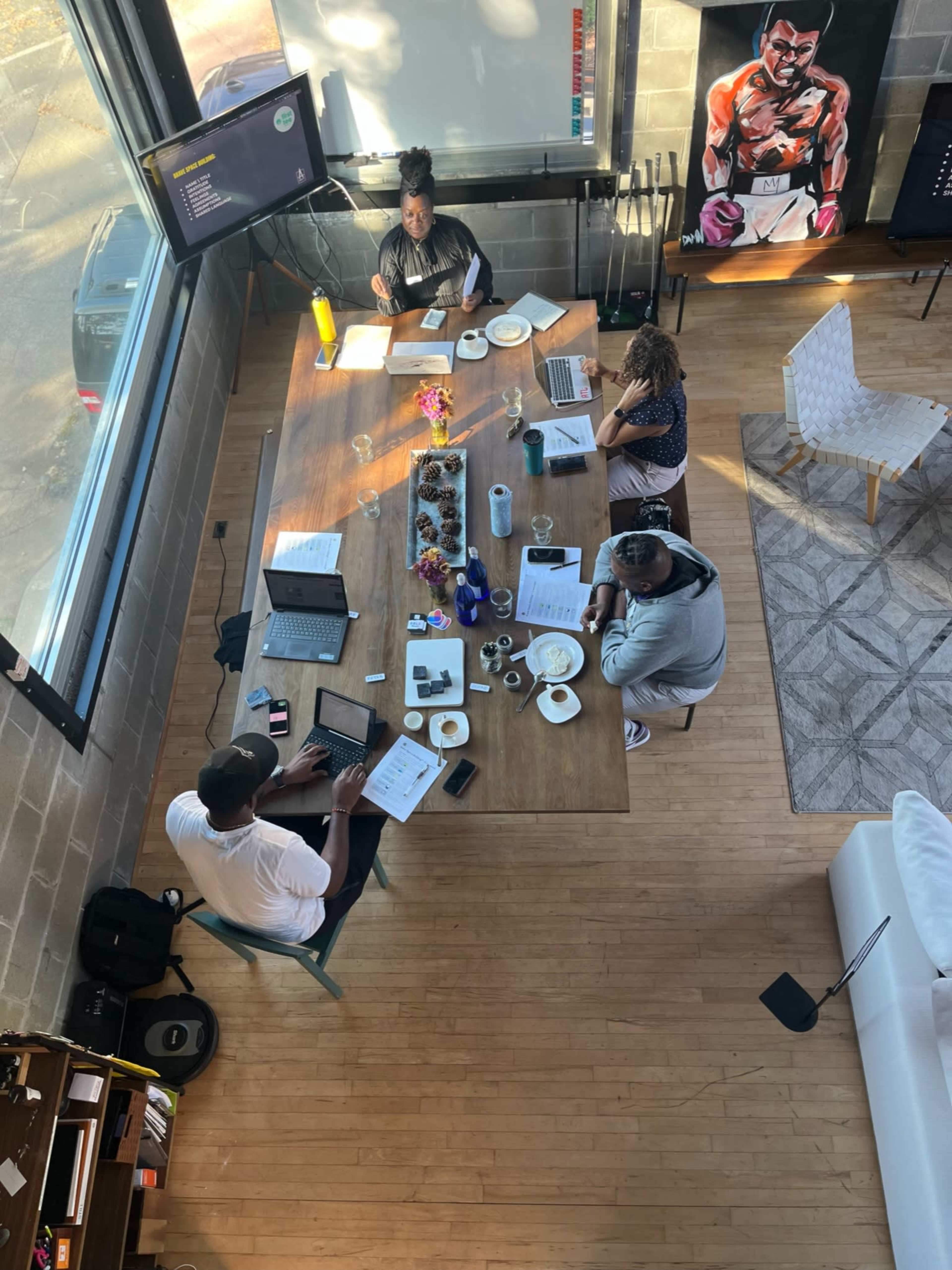 A group of four people work collaboratively around a large wooden table filled with laptops and documents in a bright room with large windows.