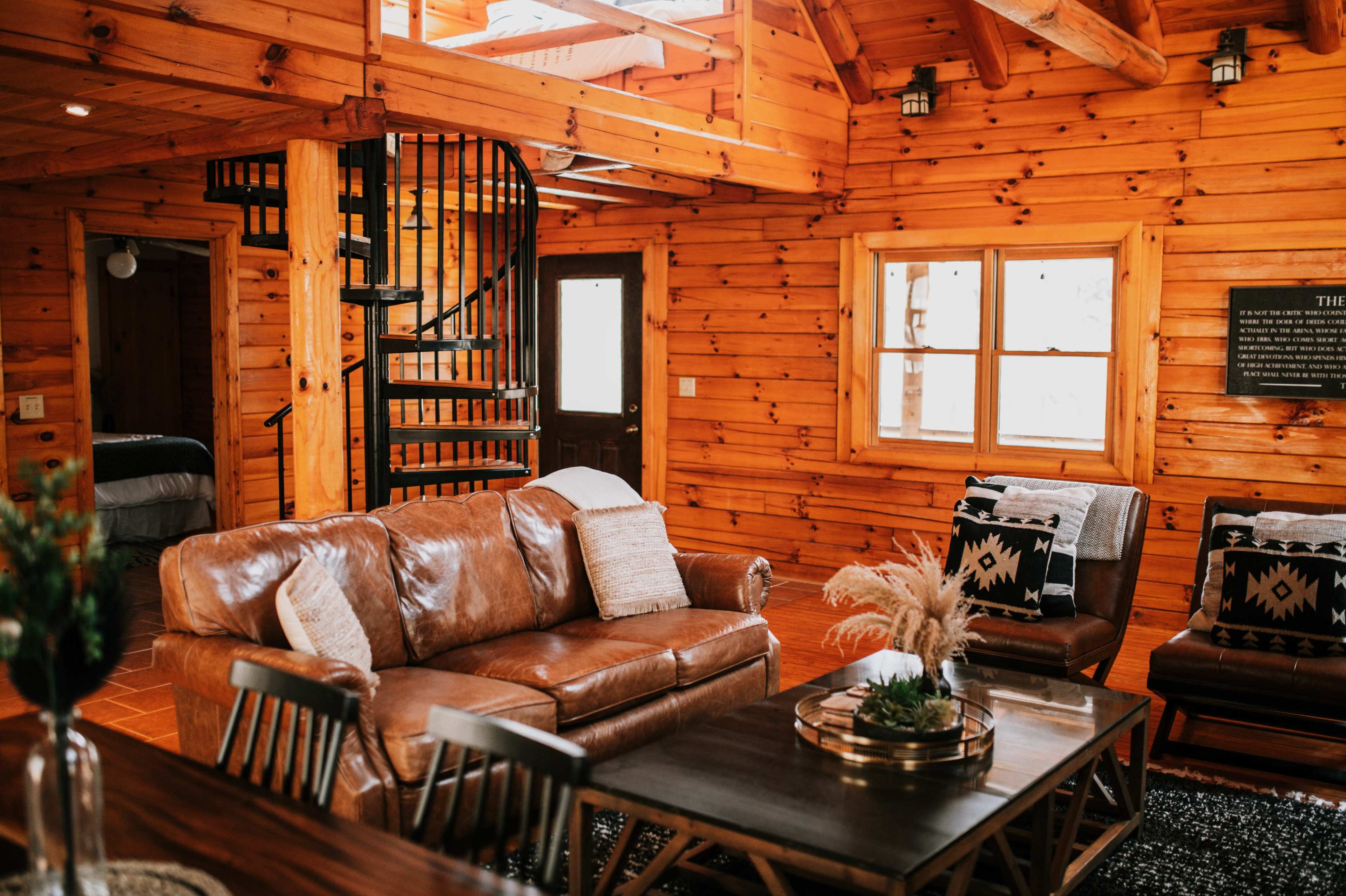 The interior of a log cabin features a cozy living area with a leather sofa, a wooden table, and a spiral staircase leading to a loft.