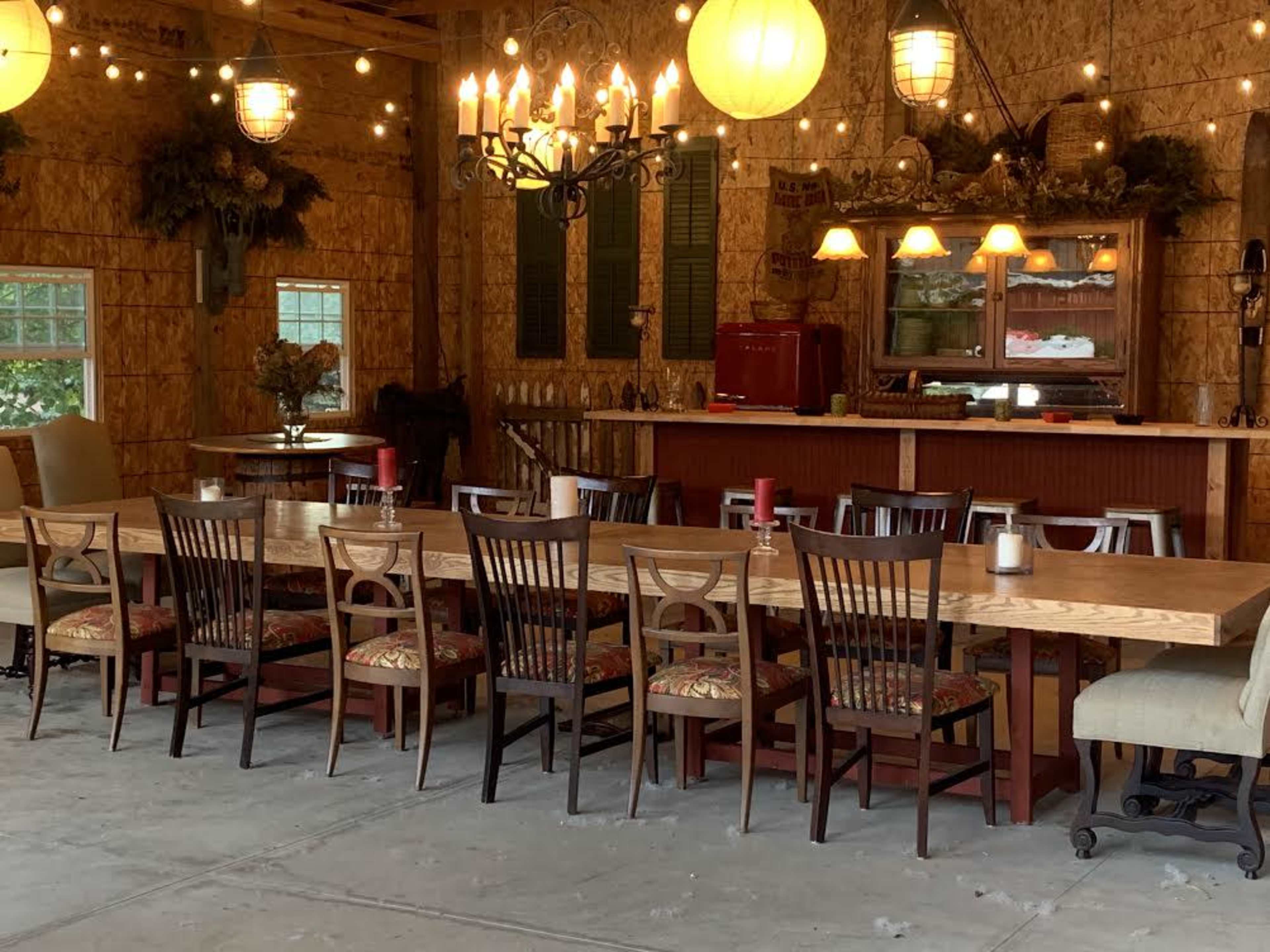 A long wooden dining table is set in a rustic room with various chairs, string lights, and a counter displaying a vintage red refrigerator.