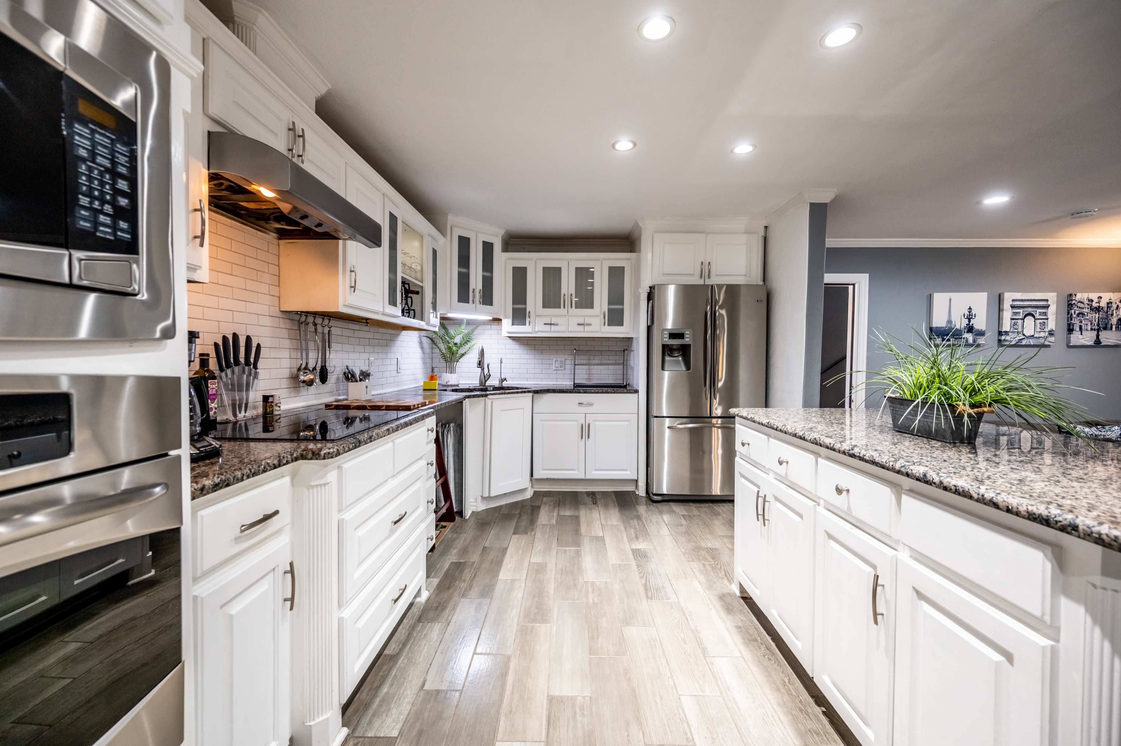 The image shows a modern kitchen with white cabinetry, stainless steel appliances, and granite countertops arranged in an open layout.