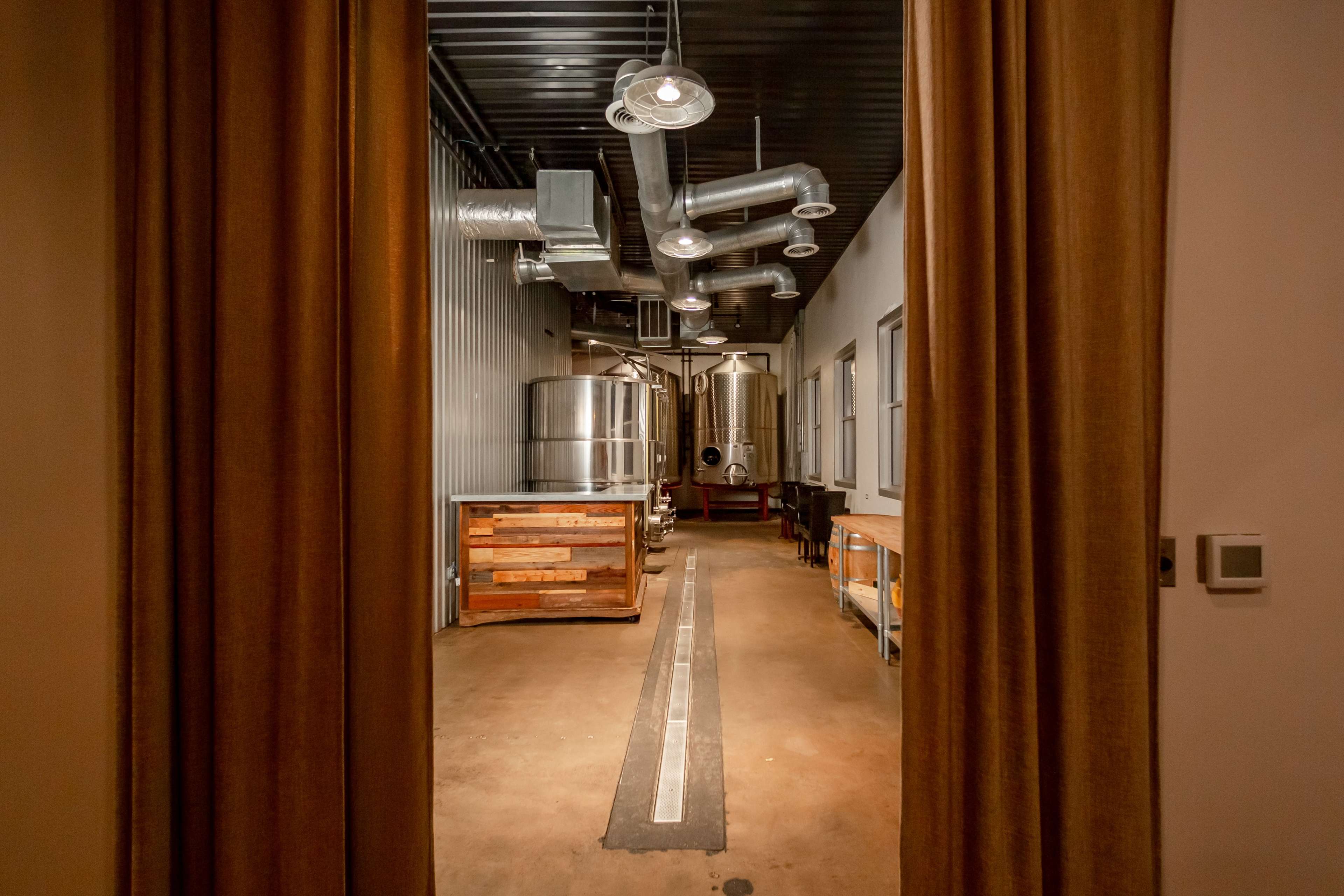 A view of a brewery interior, featuring large metal tanks, wooden tables, and industrial piping against a backdrop of muted walls.