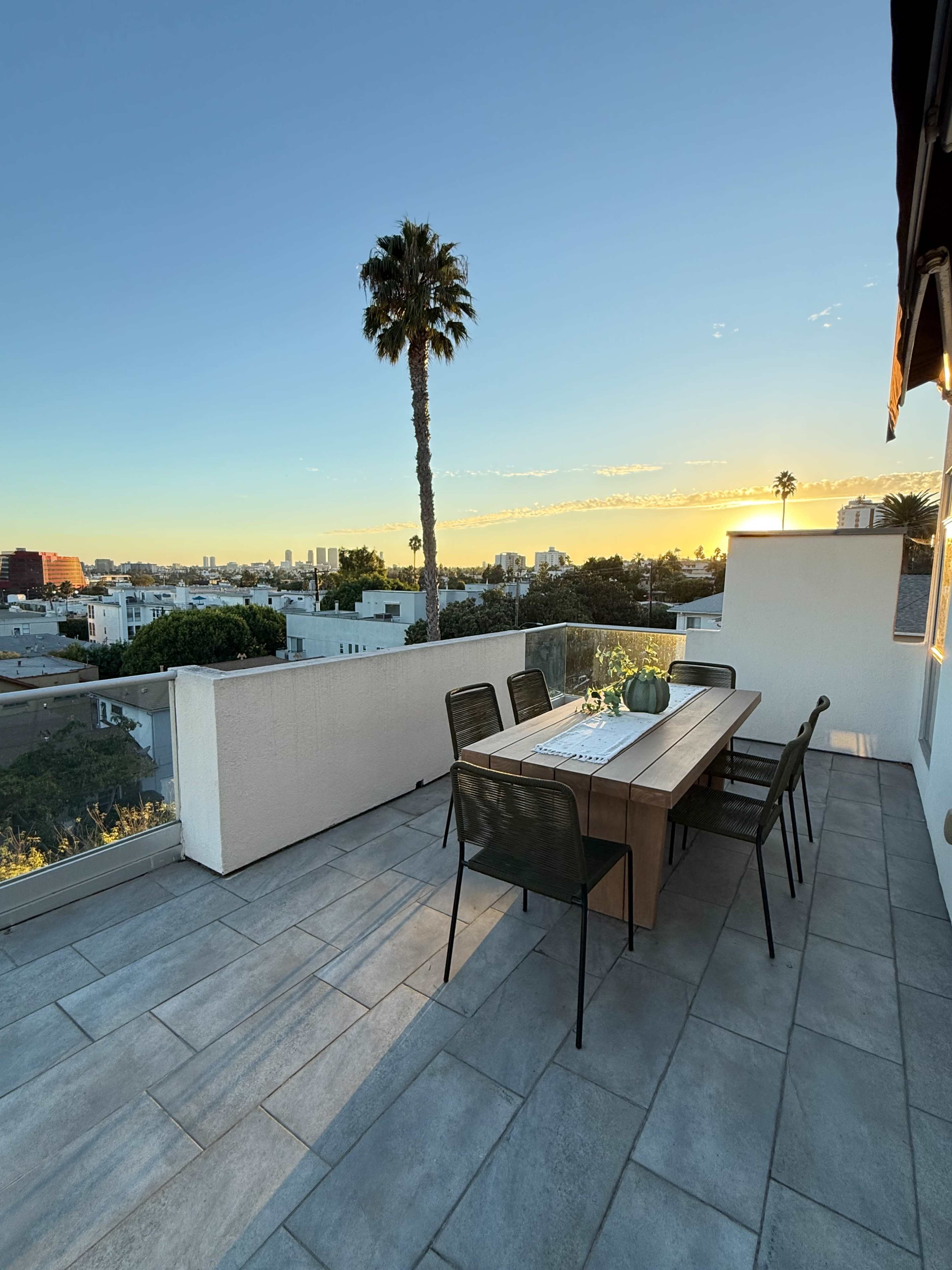 A rooftop terrace features a wooden dining table surrounded by chairs, overlooking a city skyline at sunset.
