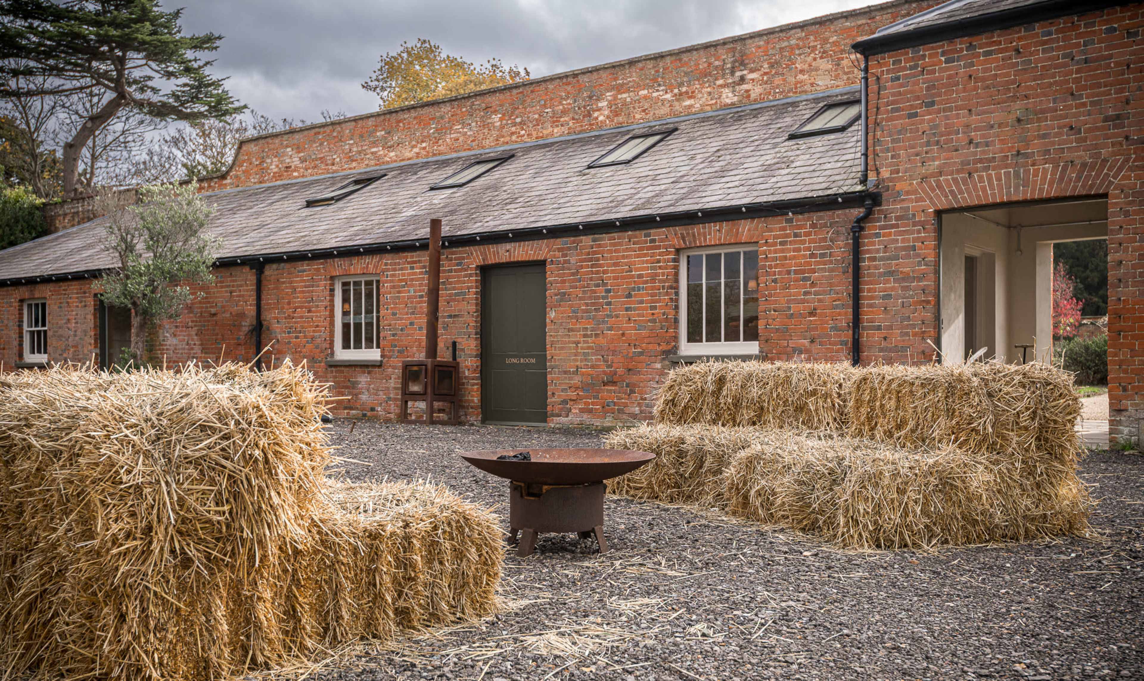 The image shows a rustic outdoor seating area made of straw bales, accompanied by a metal fire pit, set against a backdrop of a brick building with several windows.