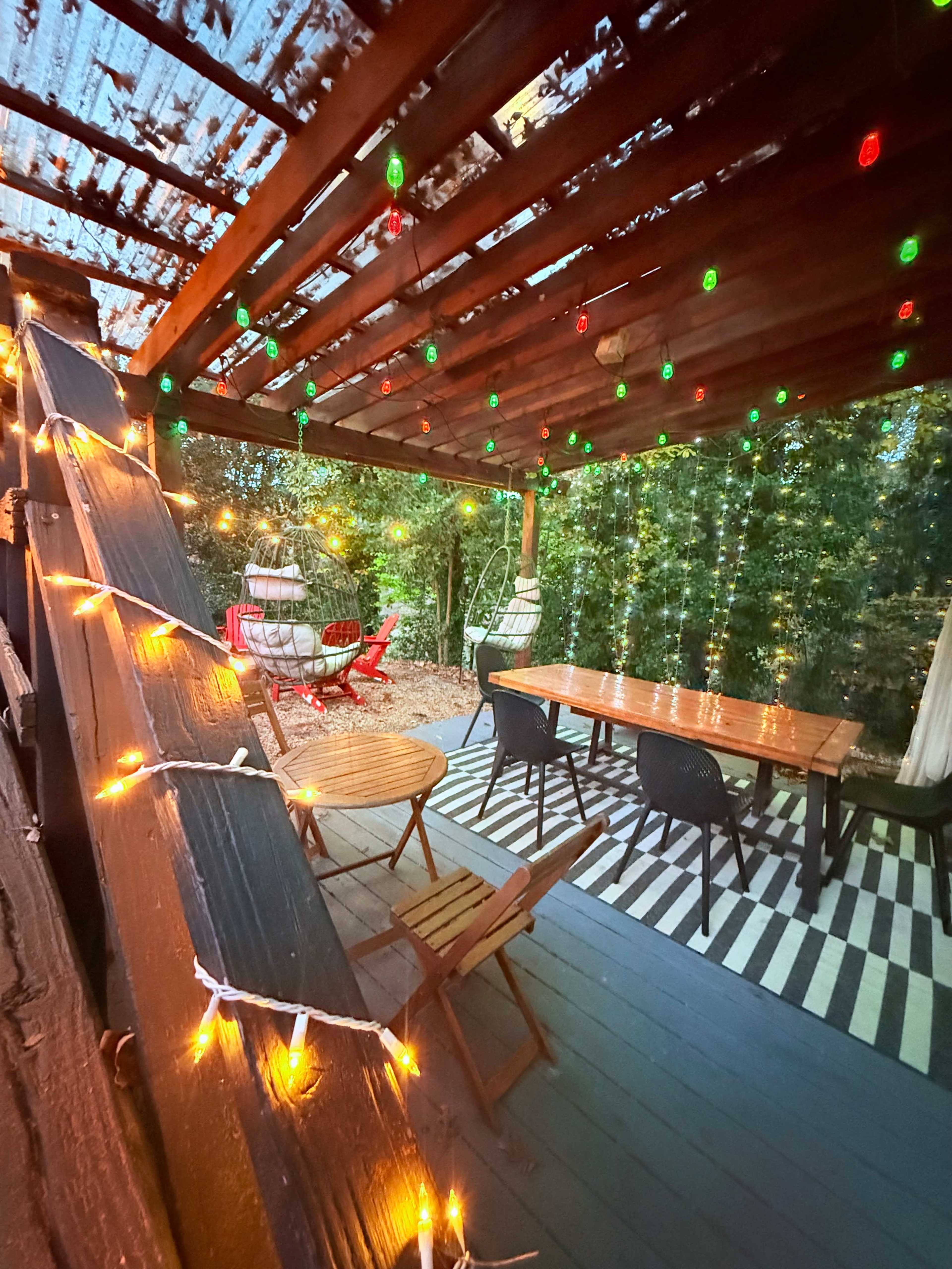 The image shows a cozy outdoor patio area with wooden furniture, decorative string lights, and a striped rug under a wooden structure.