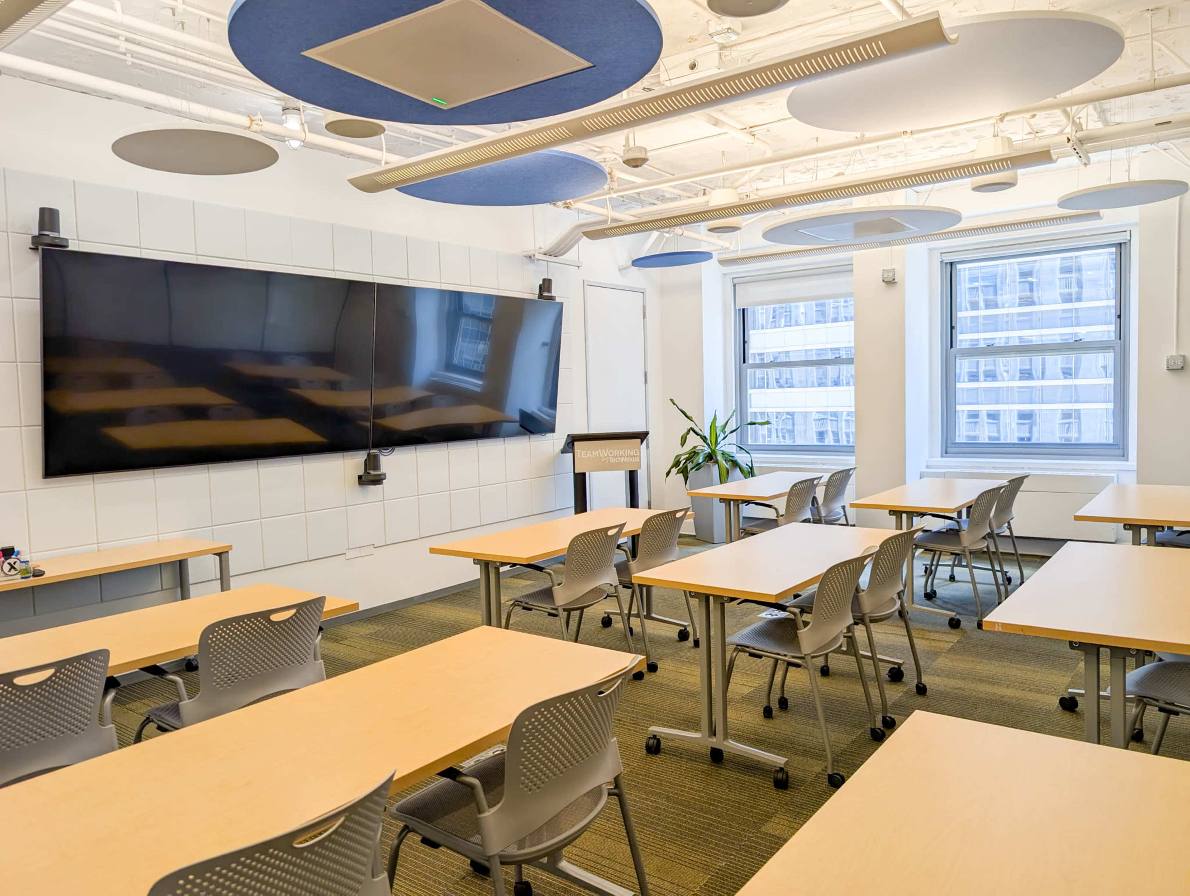The image shows a modern classroom with several tables and chairs arranged in a typical layout, featuring a large screen mounted on a wall and windows allowing natural light to enter.