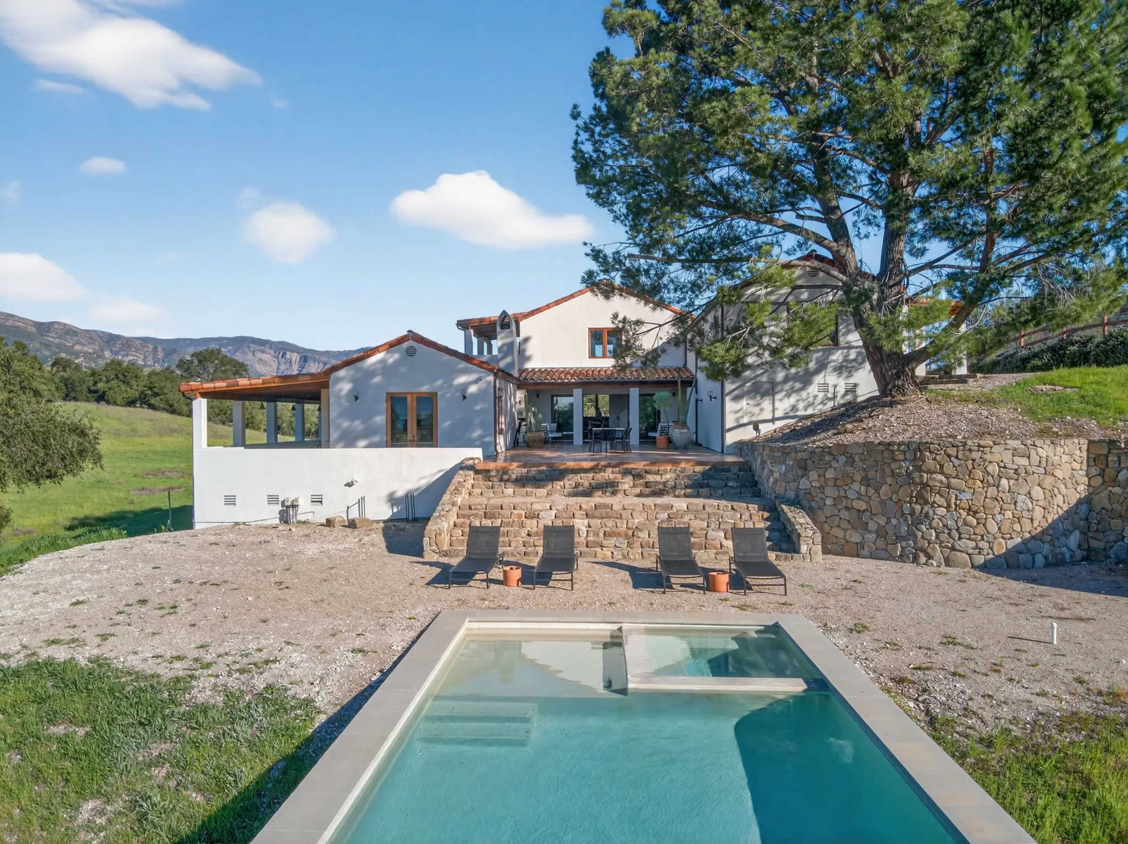 The image shows a modern house set on a hillside, featuring a swimming pool in the foreground, stone terraces, and mountainous scenery in the background.