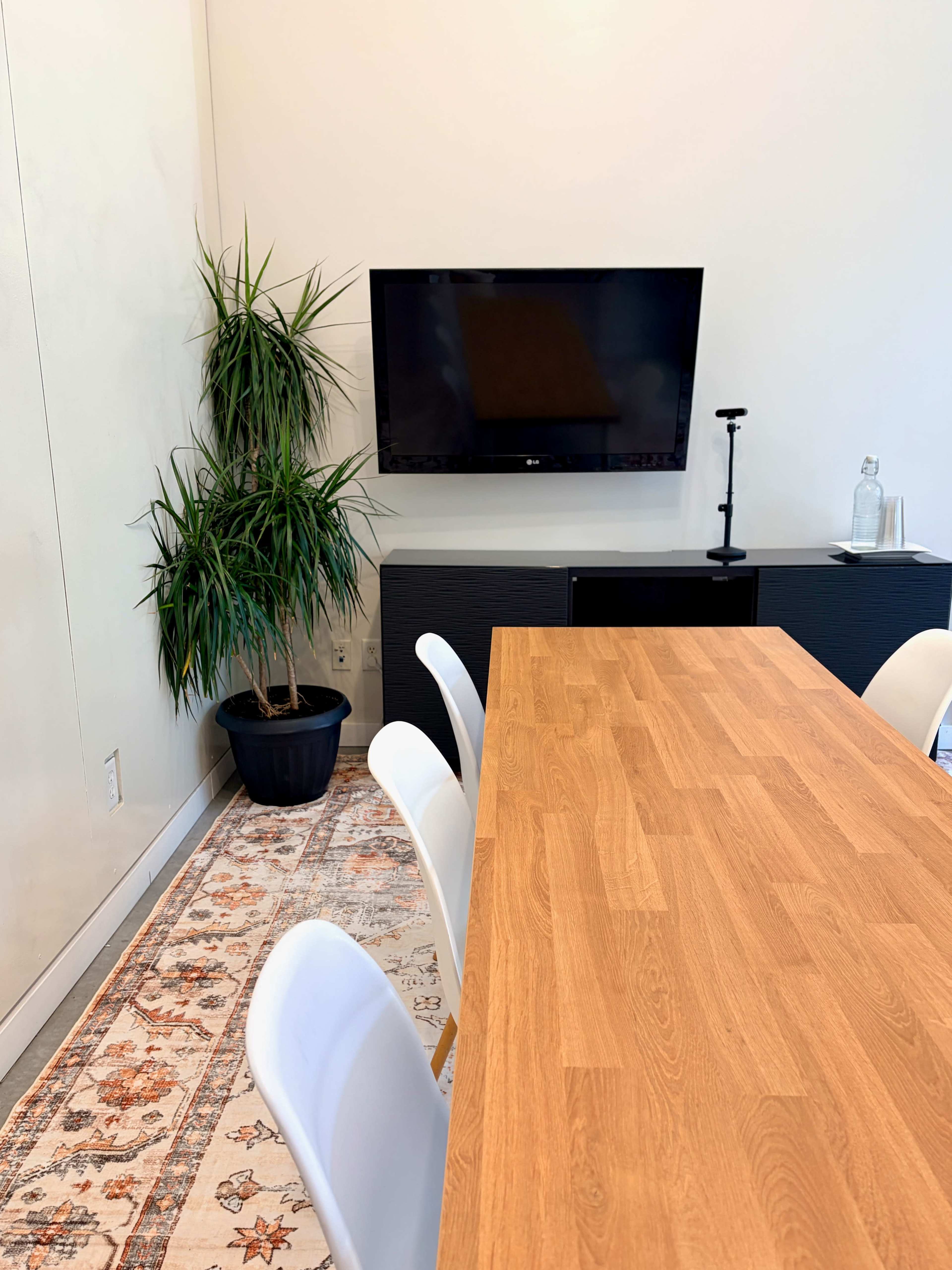 The image shows a modern conference room featuring a wooden table, white chairs, a black television mounted on the wall, a potted plant, and a decorative rug on the floor.