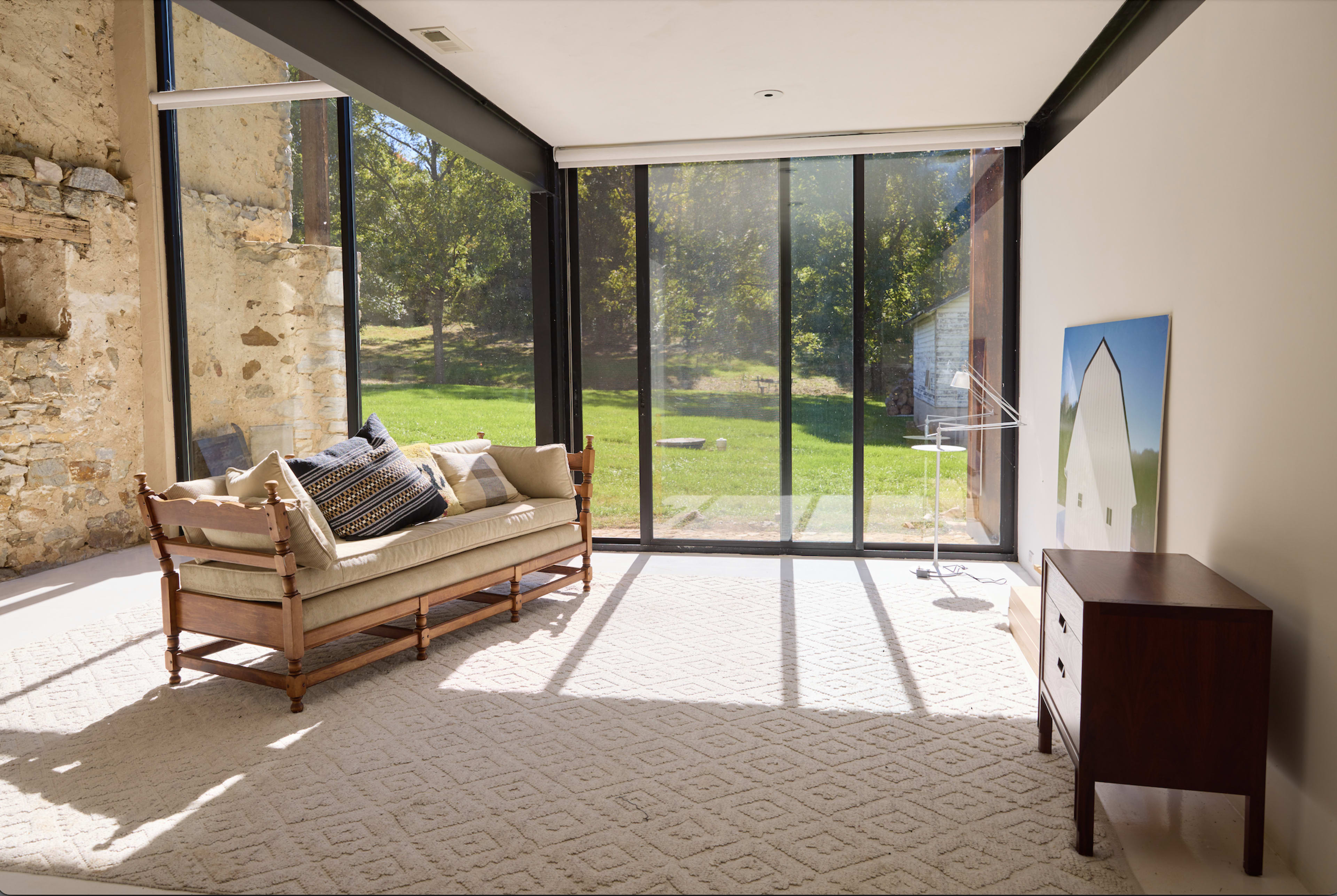 The image shows a well-lit interior with a sofa, a small wooden side table, and large glass windows overlooking a green landscape.