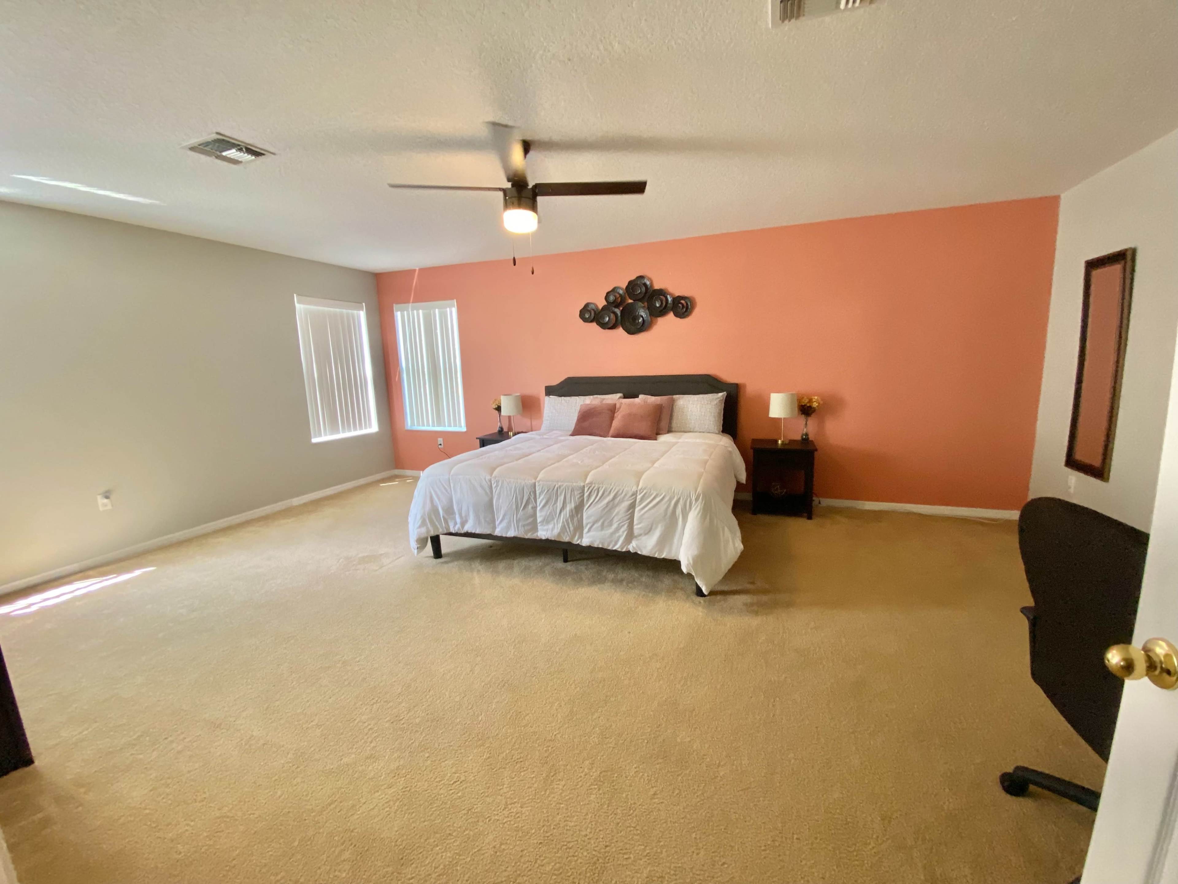 The room features a bed with white bedding against an orange accent wall, accompanied by two bedside lamps, a mirror, and a chair.