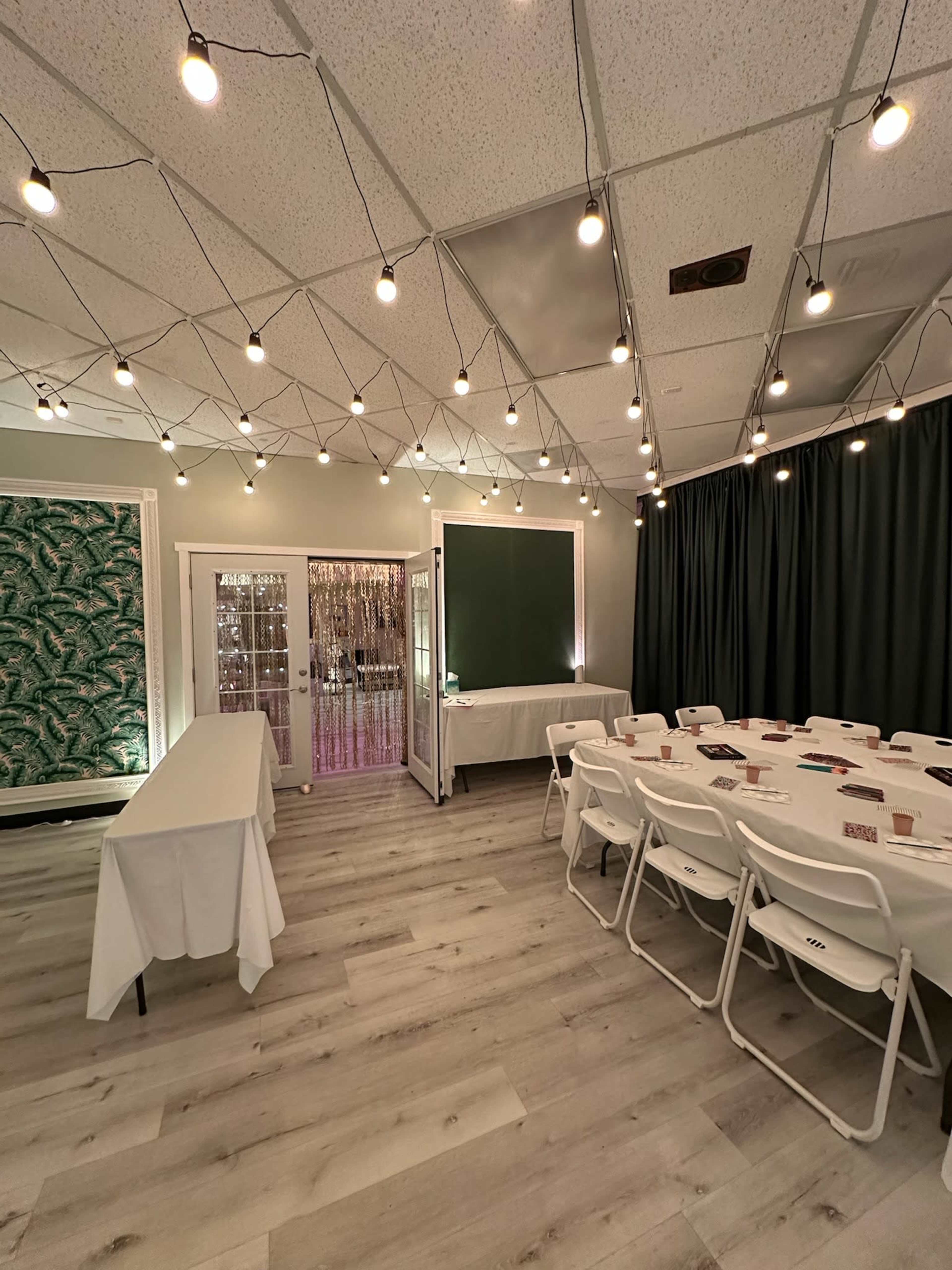 The scene shows a well-lit event space featuring a round table setup with white chairs, surrounded by a green wall design and decorative lighting overhead.