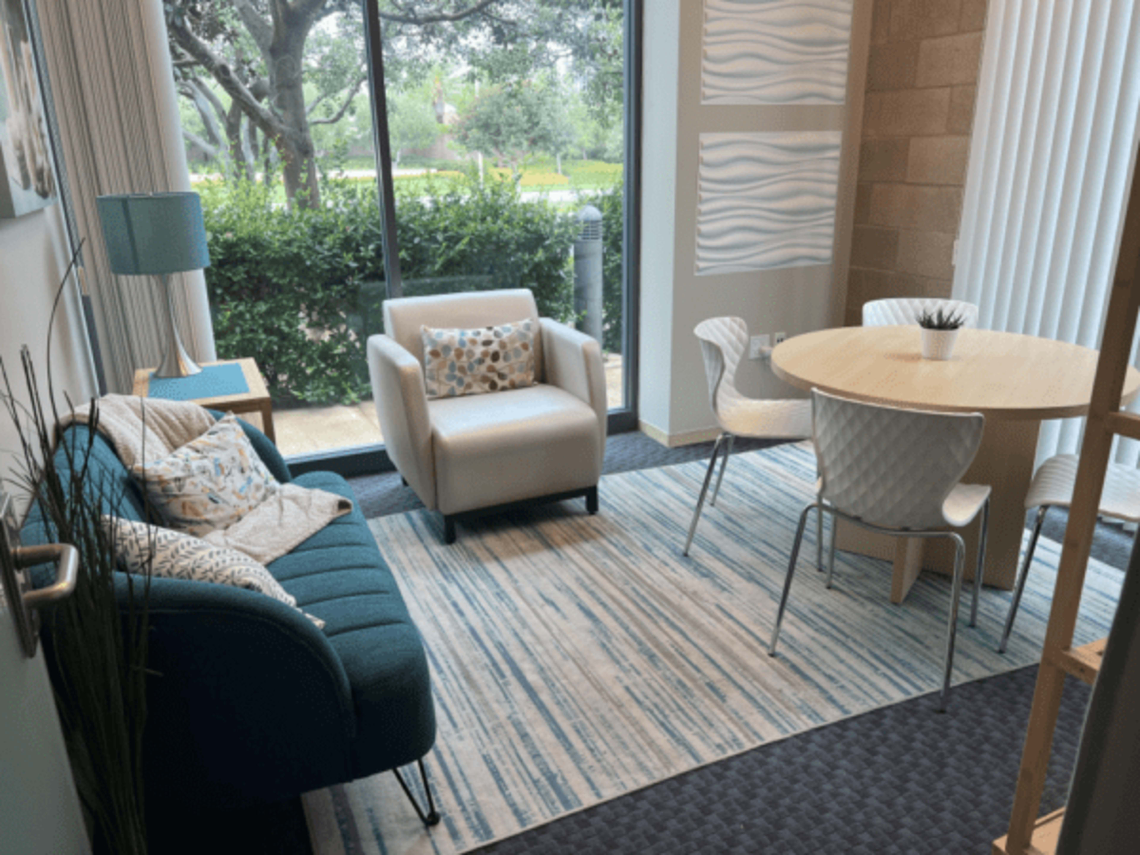 A seating area with a blue couch, two chairs, and a round table near a large window overlooking greenery.
