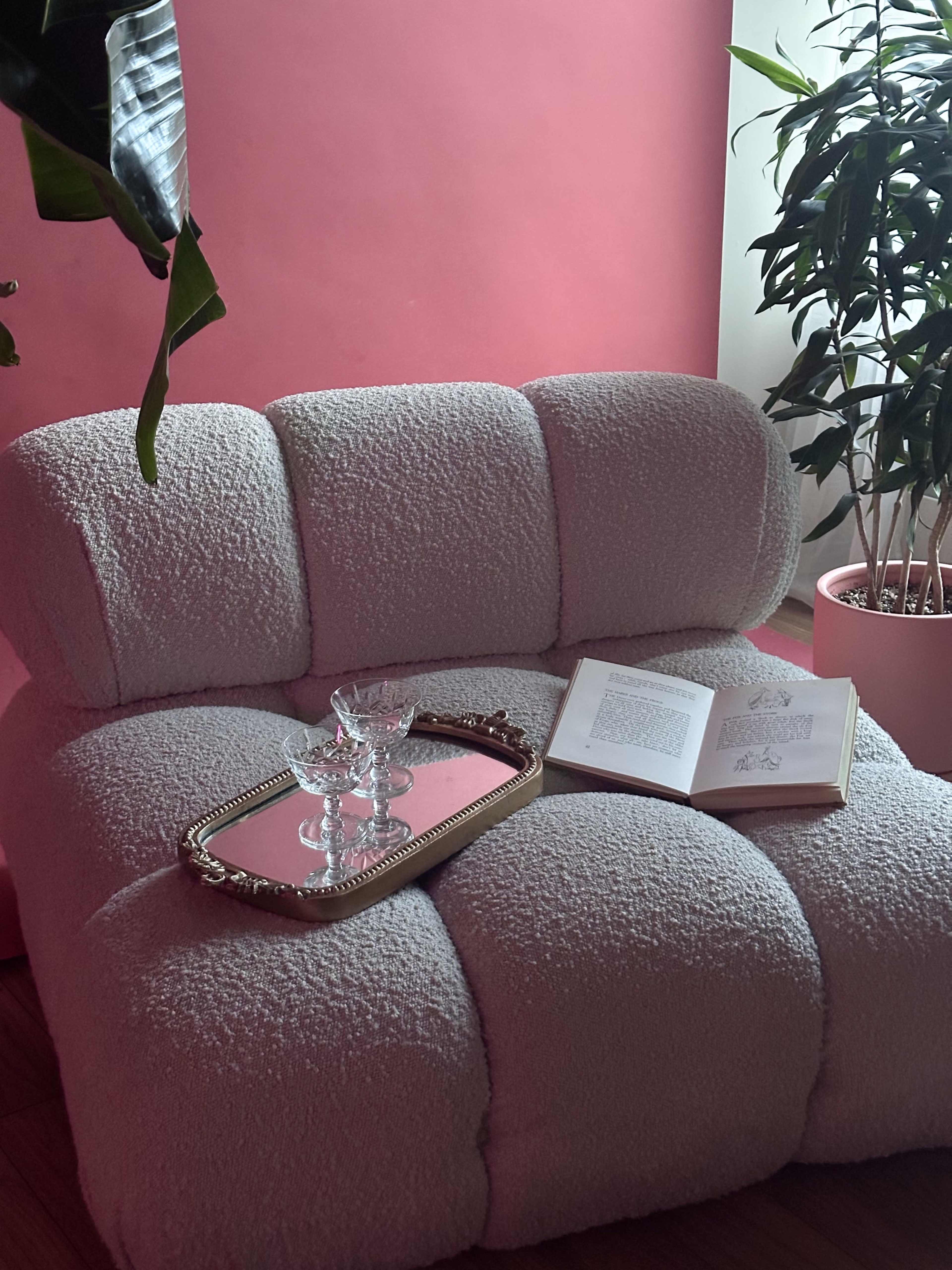 A cozy, textured pink chair is positioned beside a pink wall, adorned with a small glass dish on a tray and an open book.