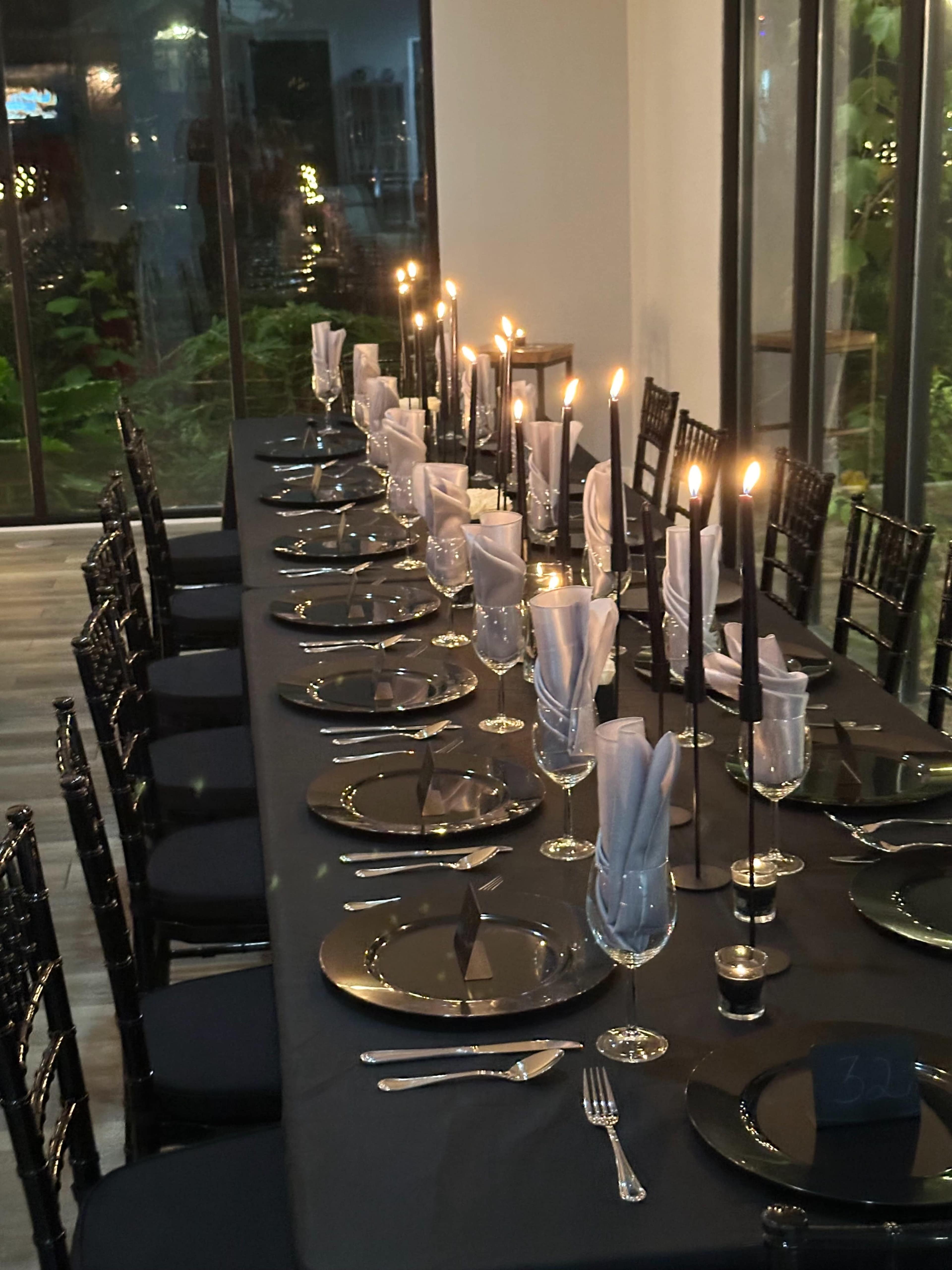 A long dining table is set with black tablecloths, elegant glassware, and lit candles along the center, surrounded by black chairs.