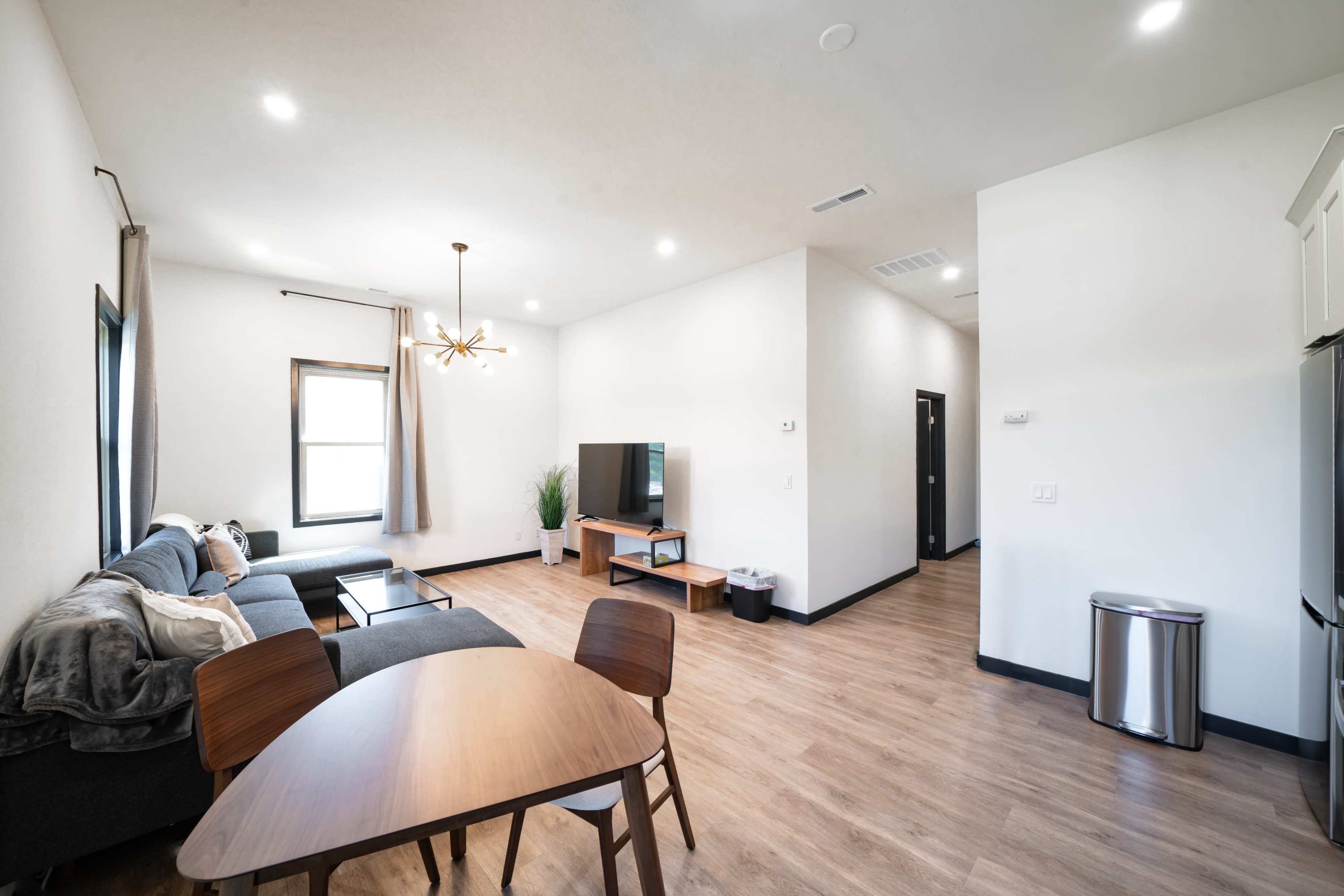 A modern, open living space featuring a gray sectional sofa, a wooden dining table, a television, and a kitchenette in a well-lit environment.