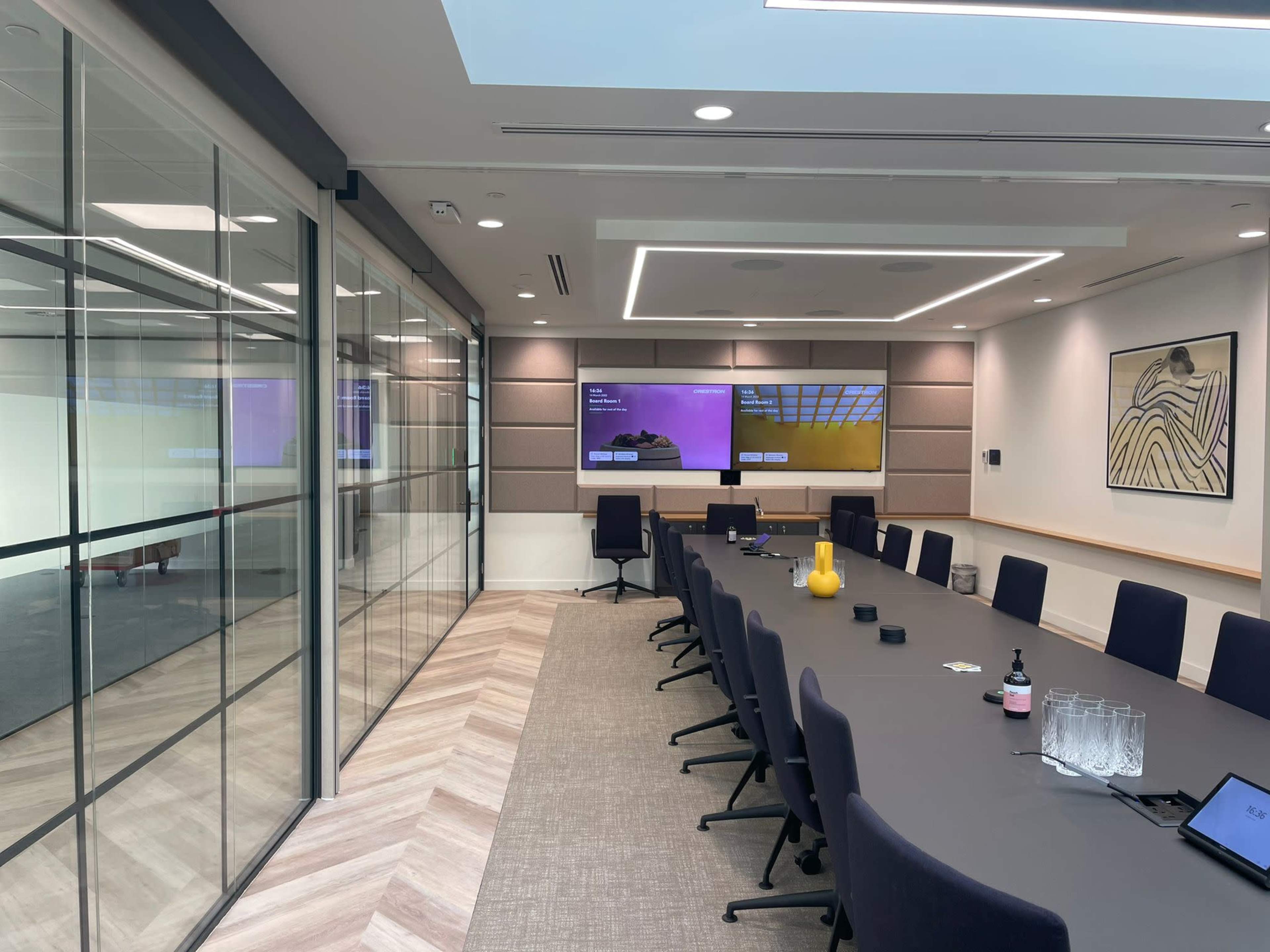 A modern conference room features a large table, high-backed chairs, two displays on the wall, and a glass partition separating it from an adjacent space.