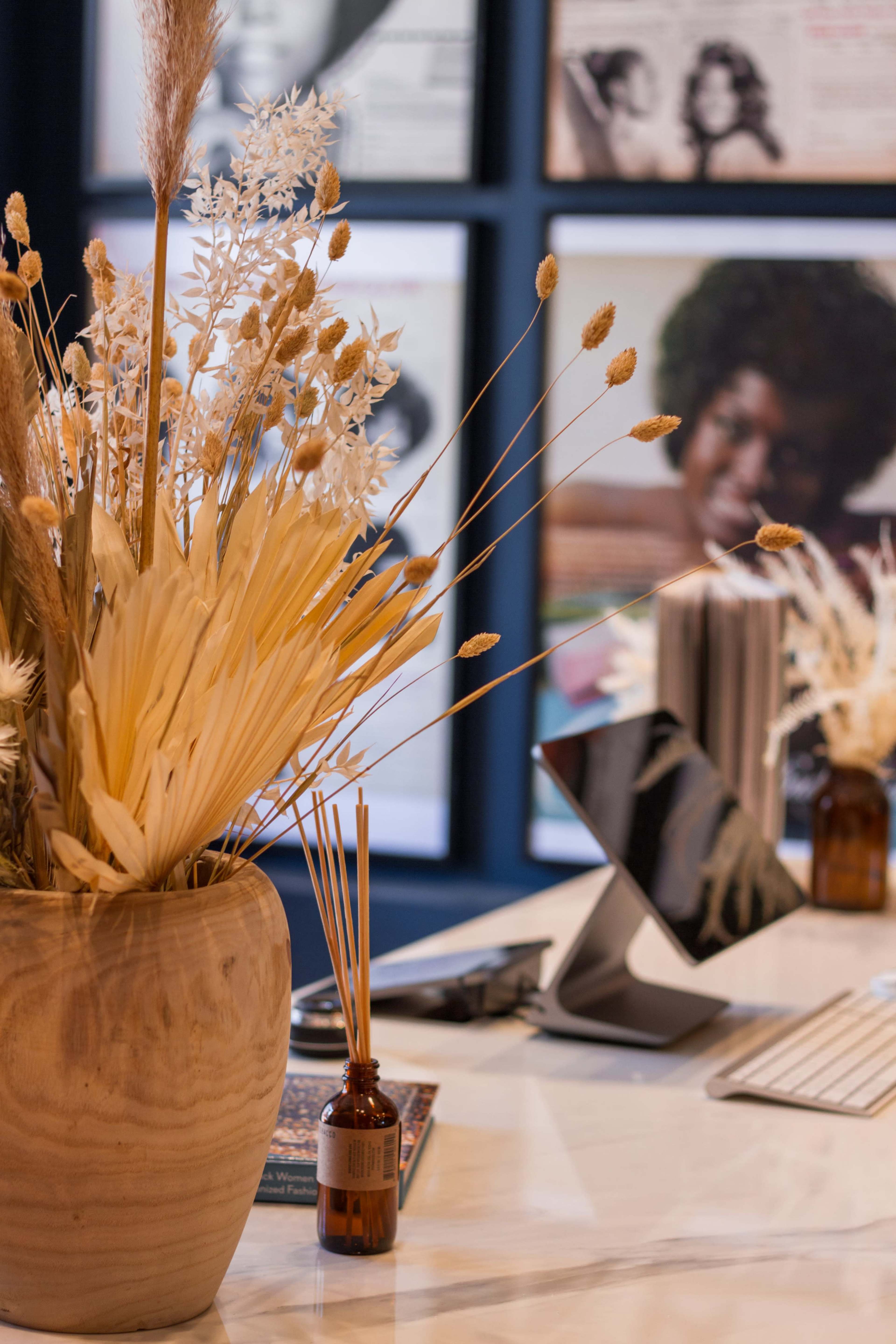 A vase of dried flowers sits on a marble desk in front of a computer and various magazines on display.