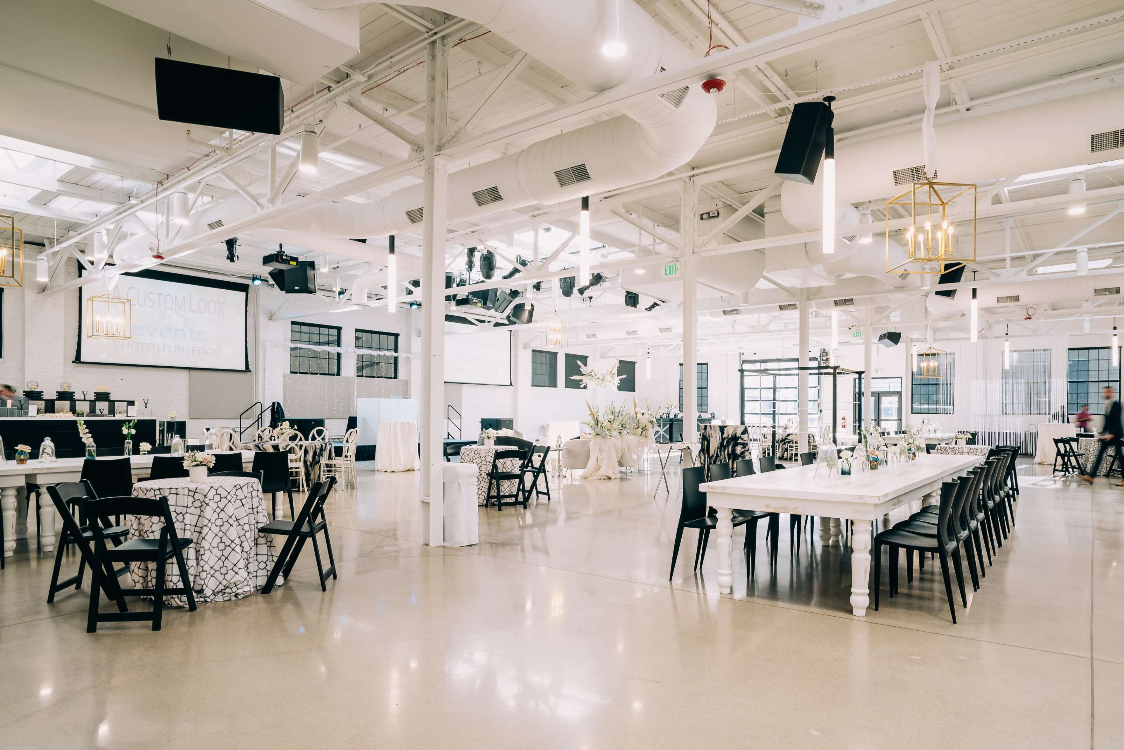 A spacious event venue features long dining tables, black folding chairs, and elegant decor, with large windows and decorative lighting.