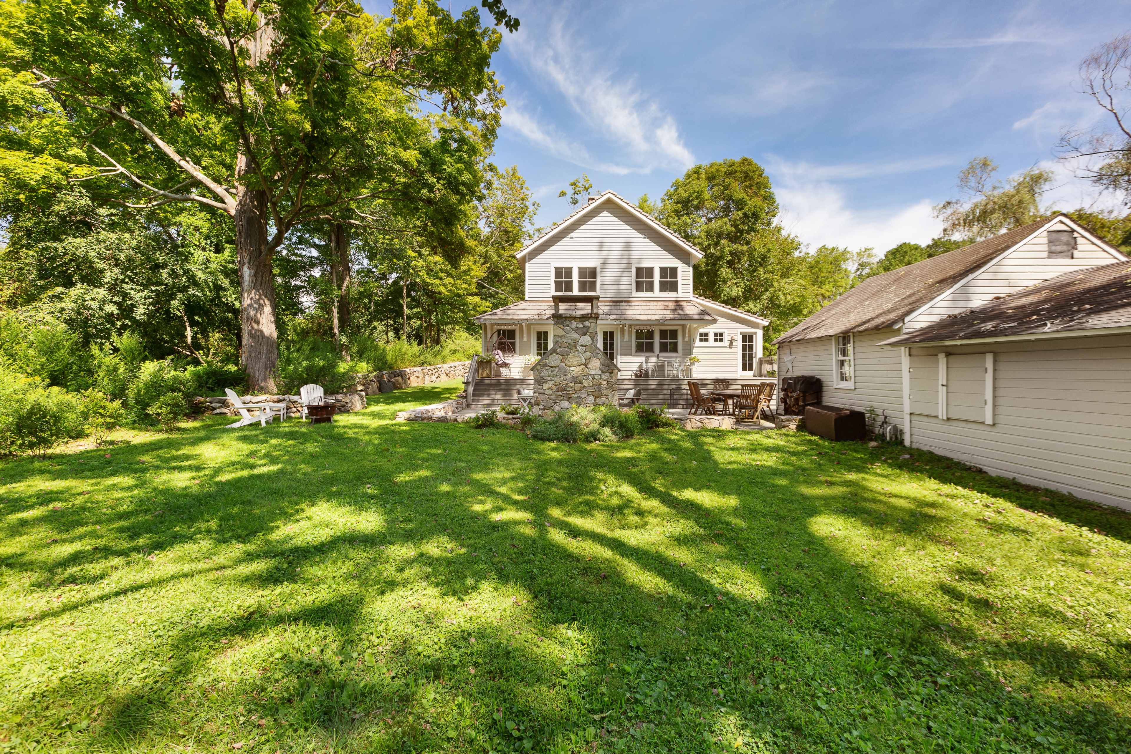 A two-story house with a deck and stone steps is situated in a grassy yard surrounded by trees.