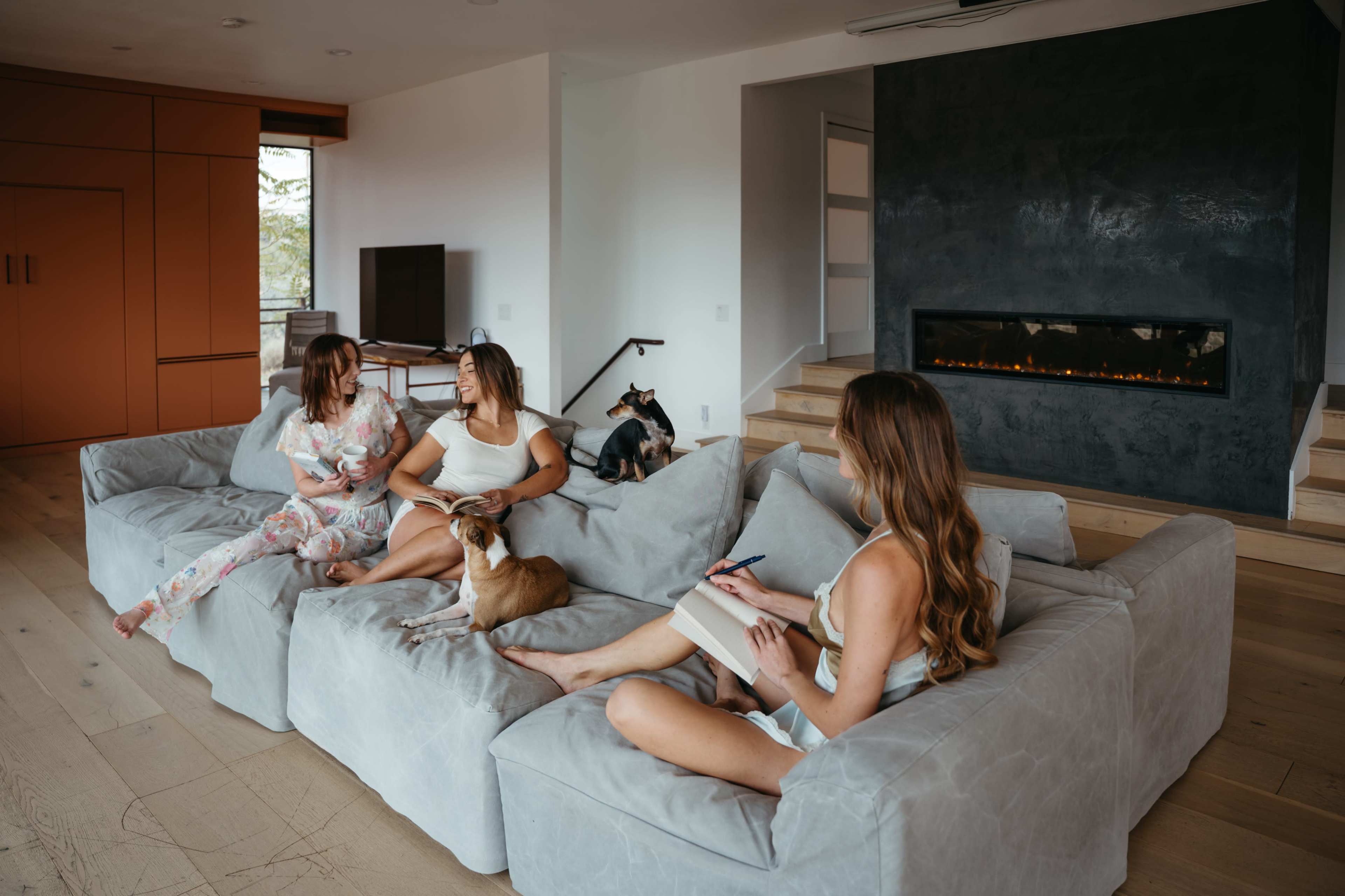 Three women are seated on a large sectional couch in a modern living room, with two dogs lounging nearby while one woman writes in a notebook.