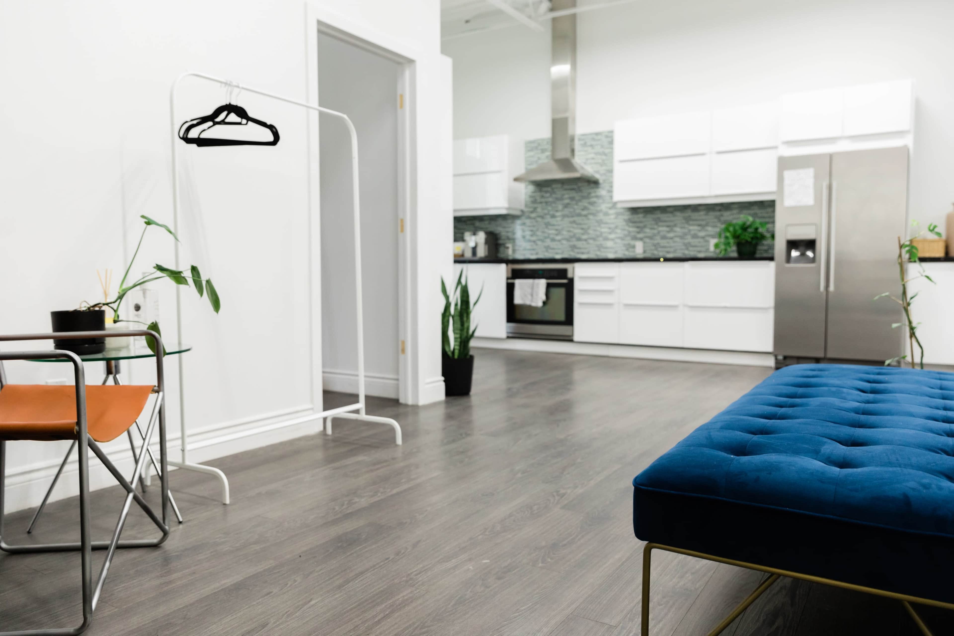A modern kitchen and living area feature a plush blue bench, a small table with an orange chair, and a clothing rack with plants in the background.