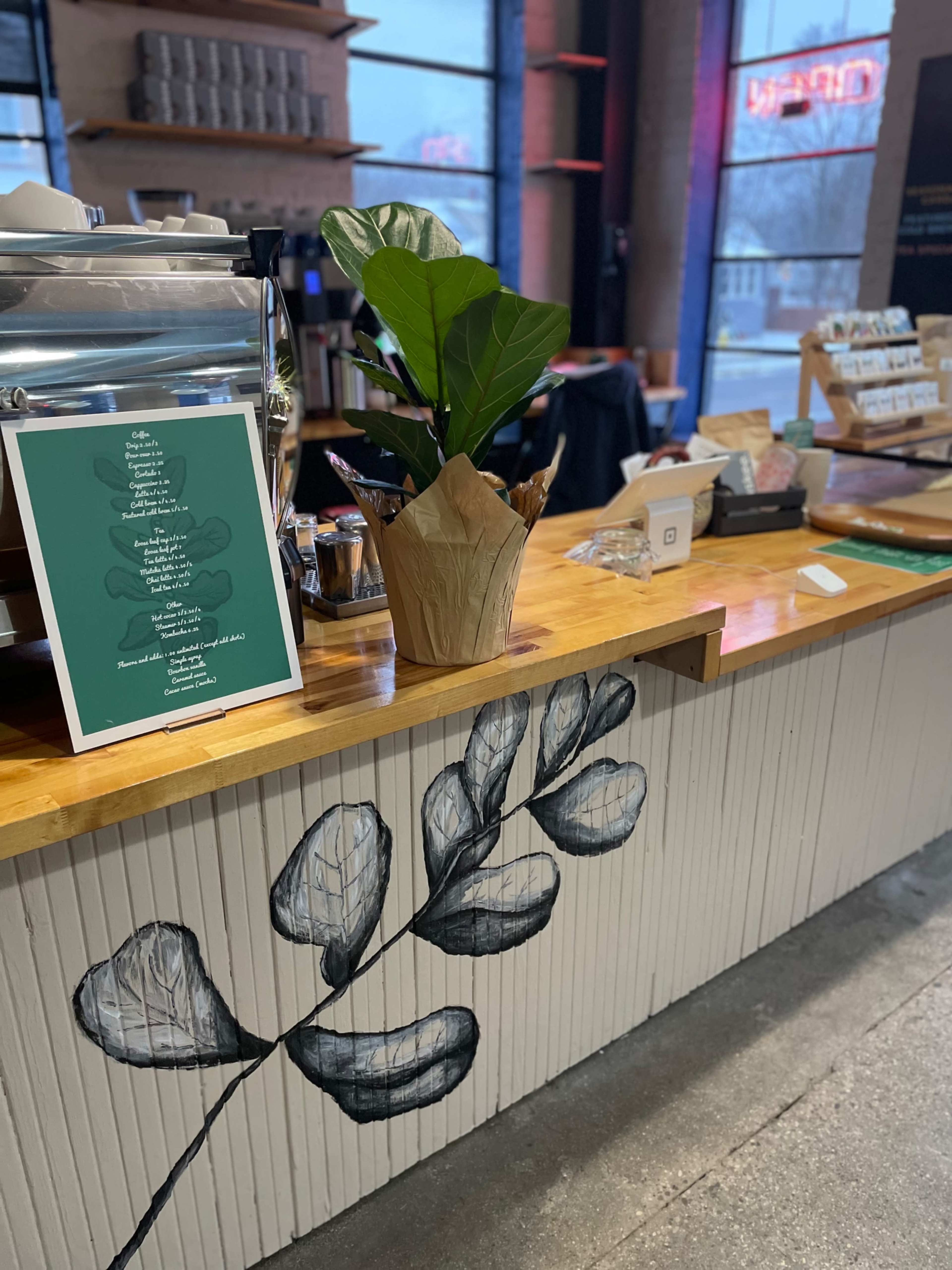 A coffee shop counter featuring a menu on a green board, a potted plant, and a wall with a leaf mural.
