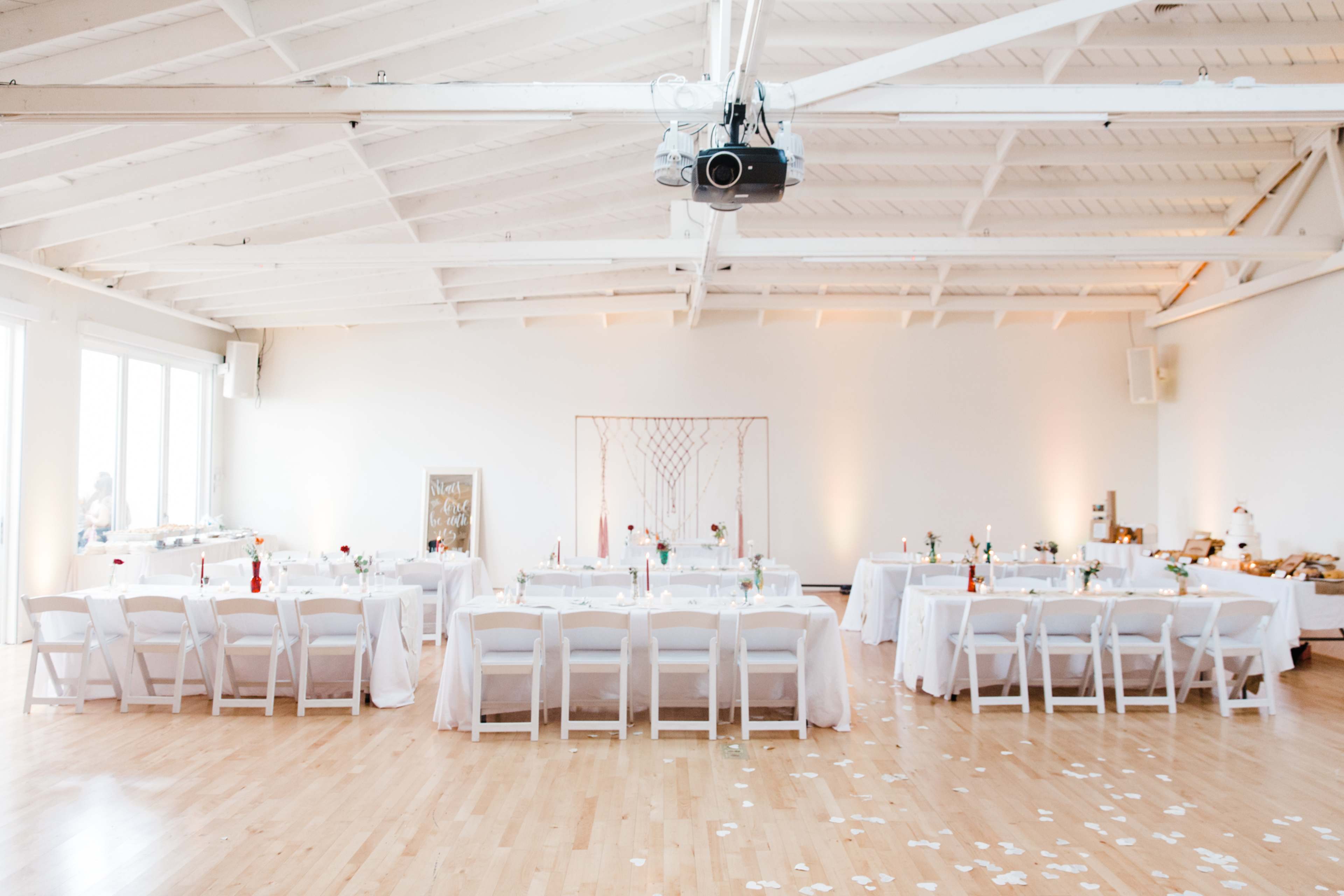 A large banquet hall is set for an event, featuring multiple tables covered with white linens, arranged with centerpieces and scattered petals, under soft lighting.