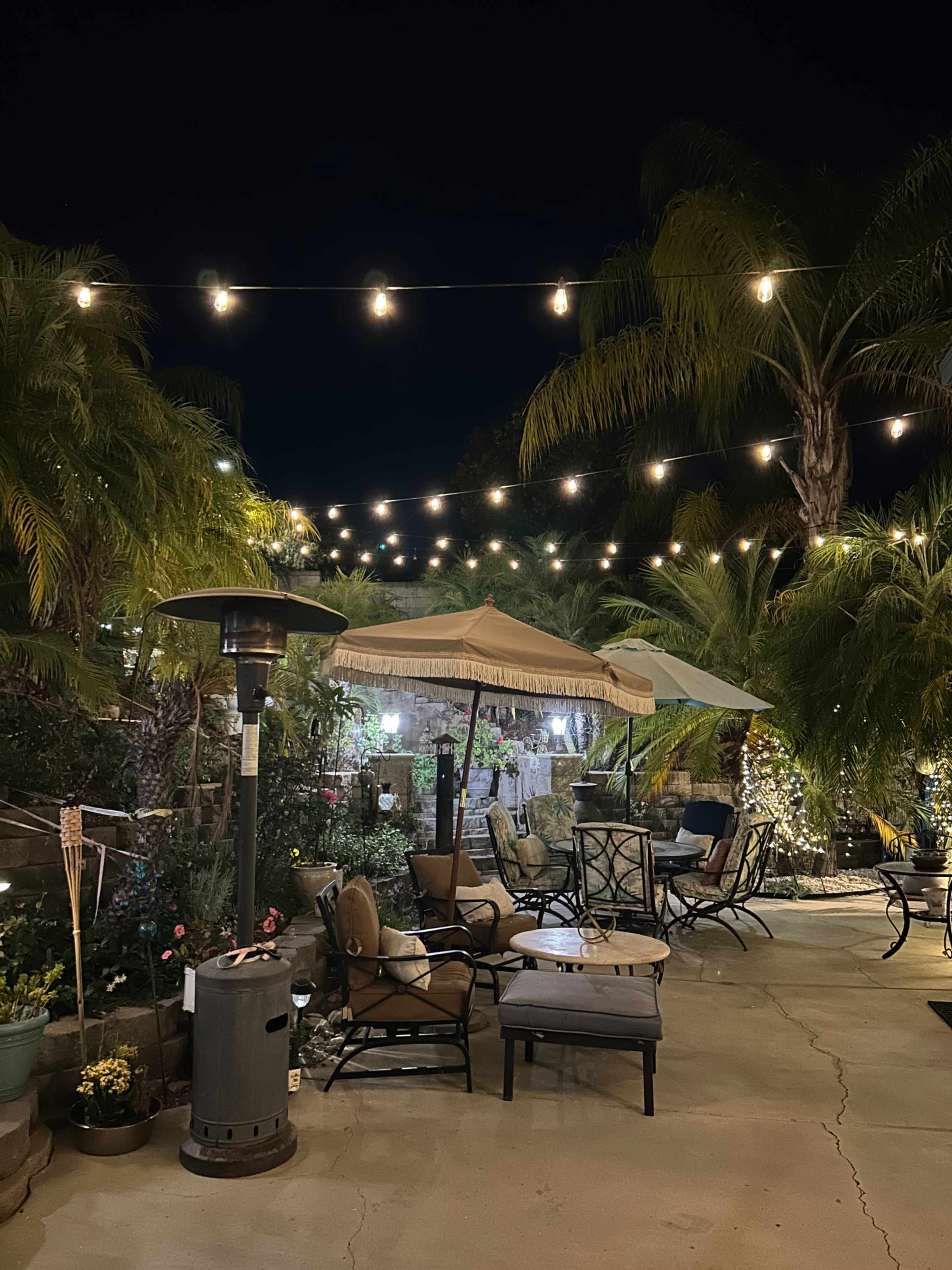 A cozy outdoor patio area is illuminated by string lights, featuring a gazebo, comfortable seating, and lush palm trees.