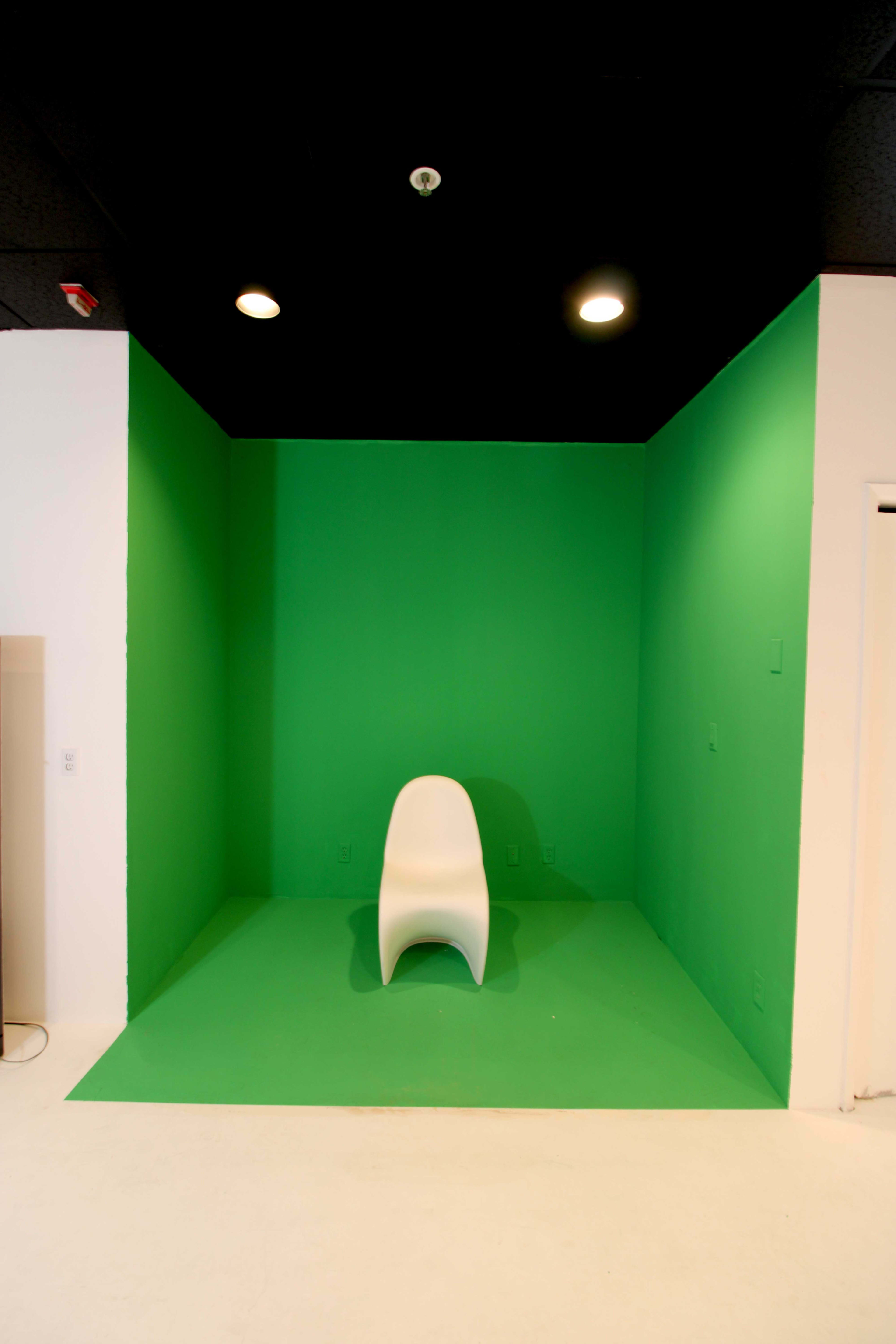 The image shows a green-painted corner with a white chair positioned in the center, under overhead lights.