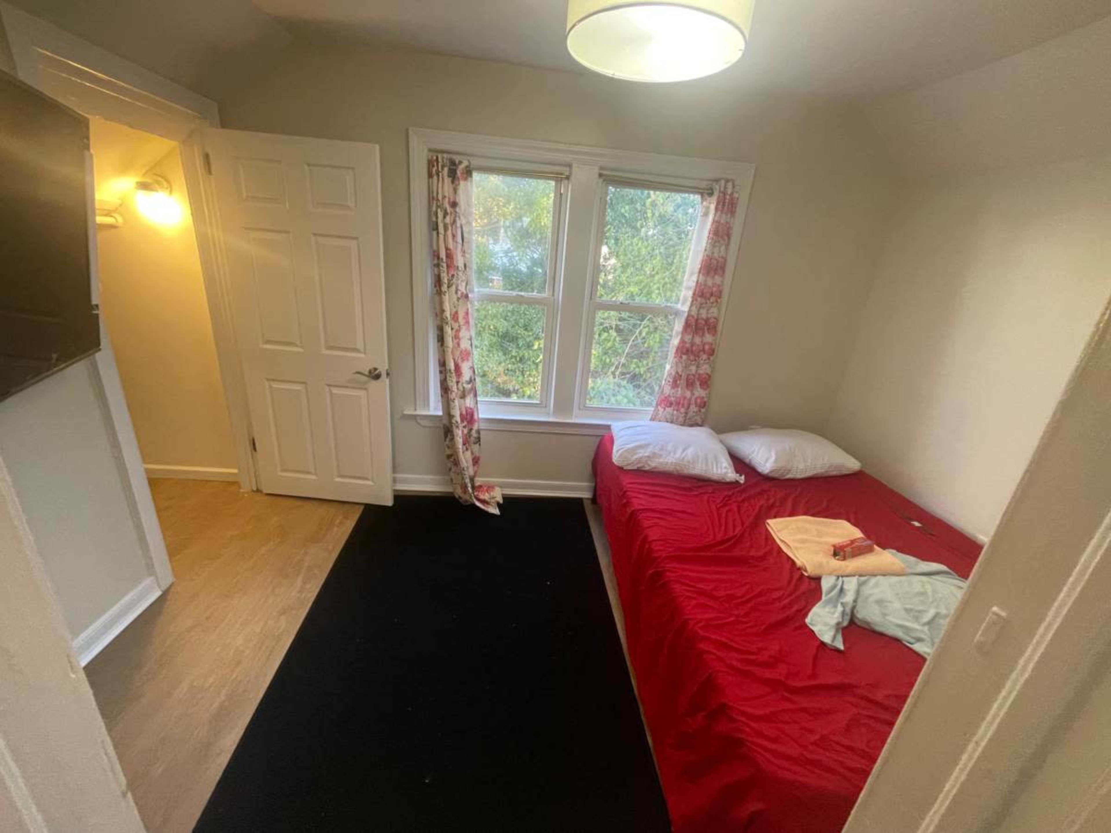 The image shows a small bedroom with a red bedspread, two pillows, and a doorway leading to another room.