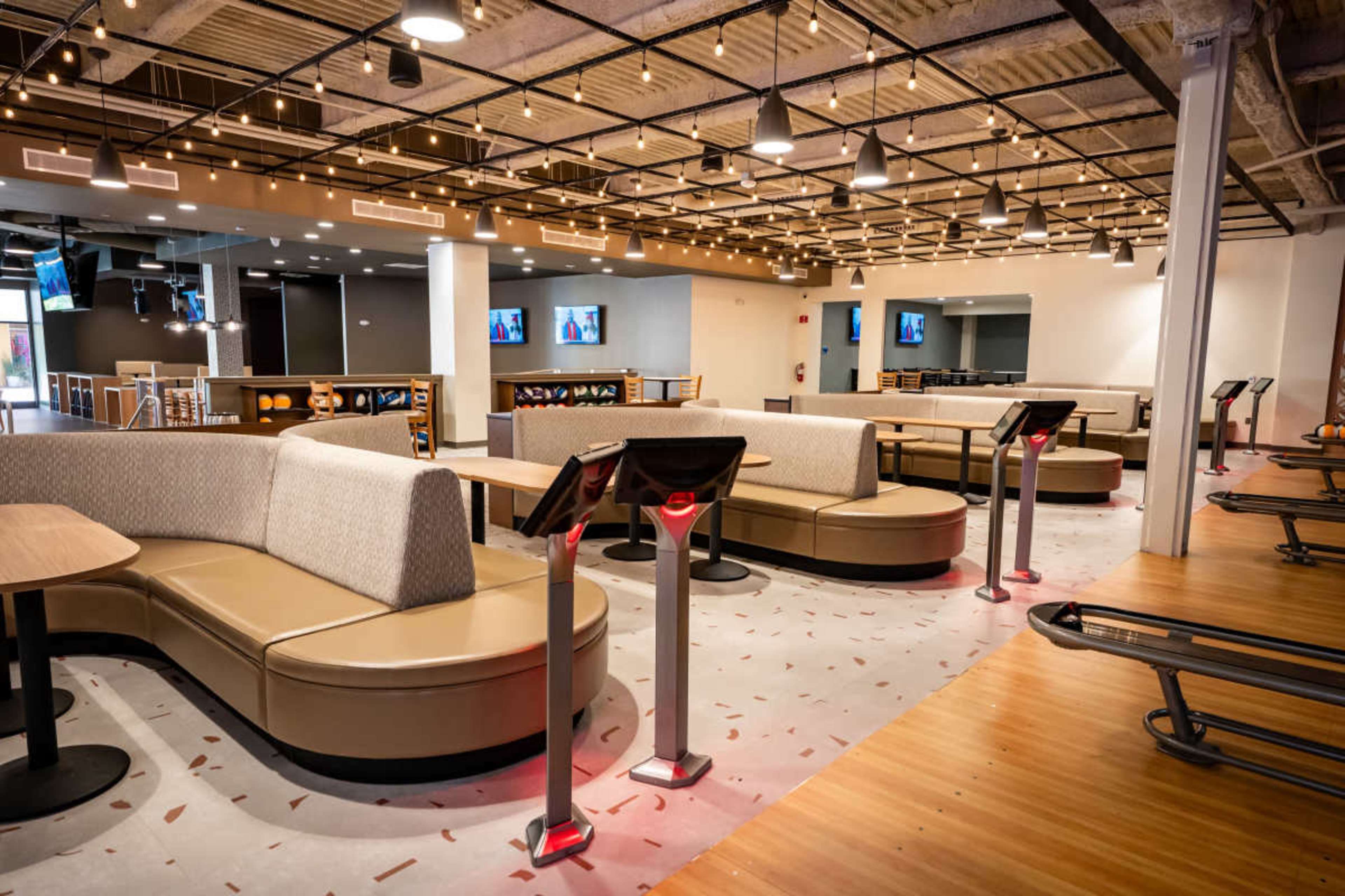 The image shows a modern bowling alley interior featuring curved seating areas, multiple bowling lanes, and screens displaying scores.