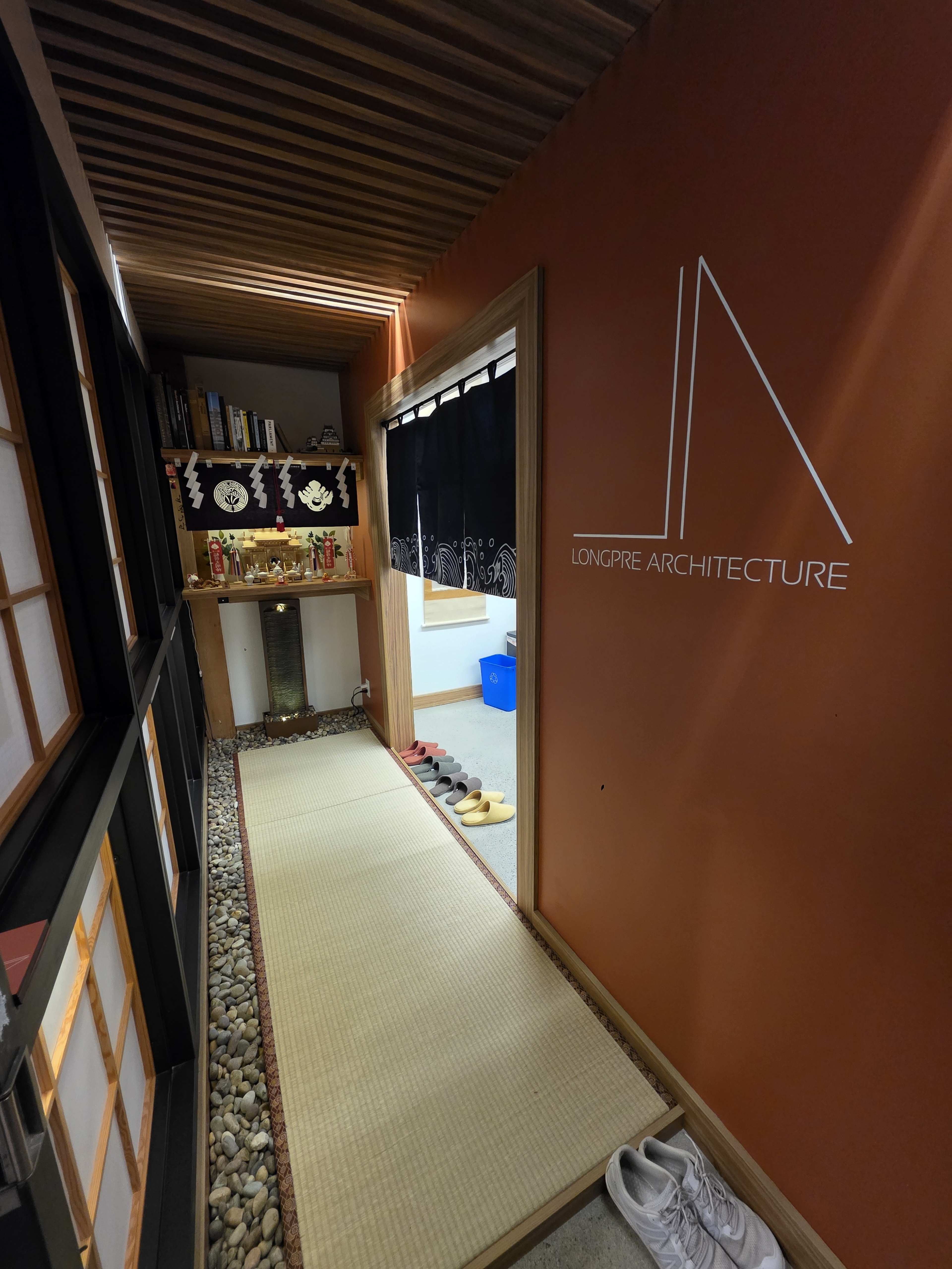 The image shows a narrow hallway with tatami flooring, lined with a shelving unit on one side and a dark curtain on the other, featuring the logo of "LONGPRE ARCHITECTURE" on an orange wall.