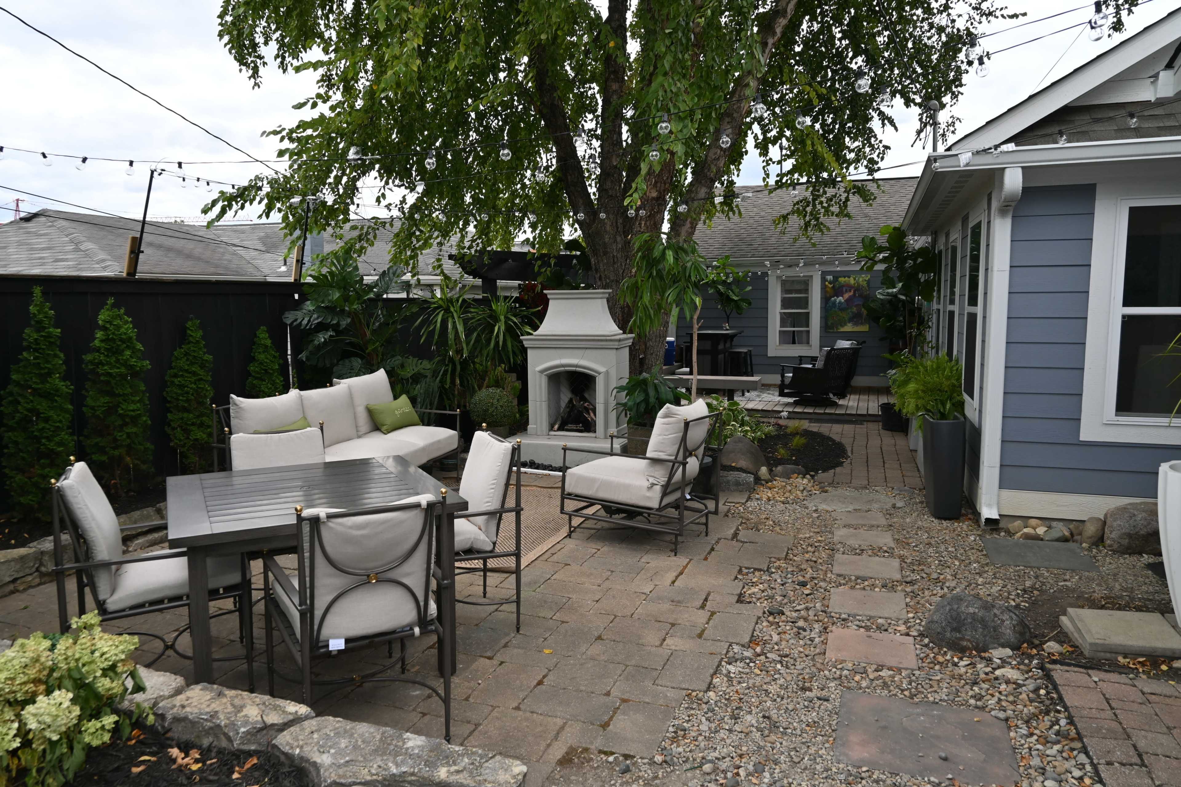 A landscaped backyard features a seating area with cushioned chairs and a table, a fireplace, and surrounding greenery.