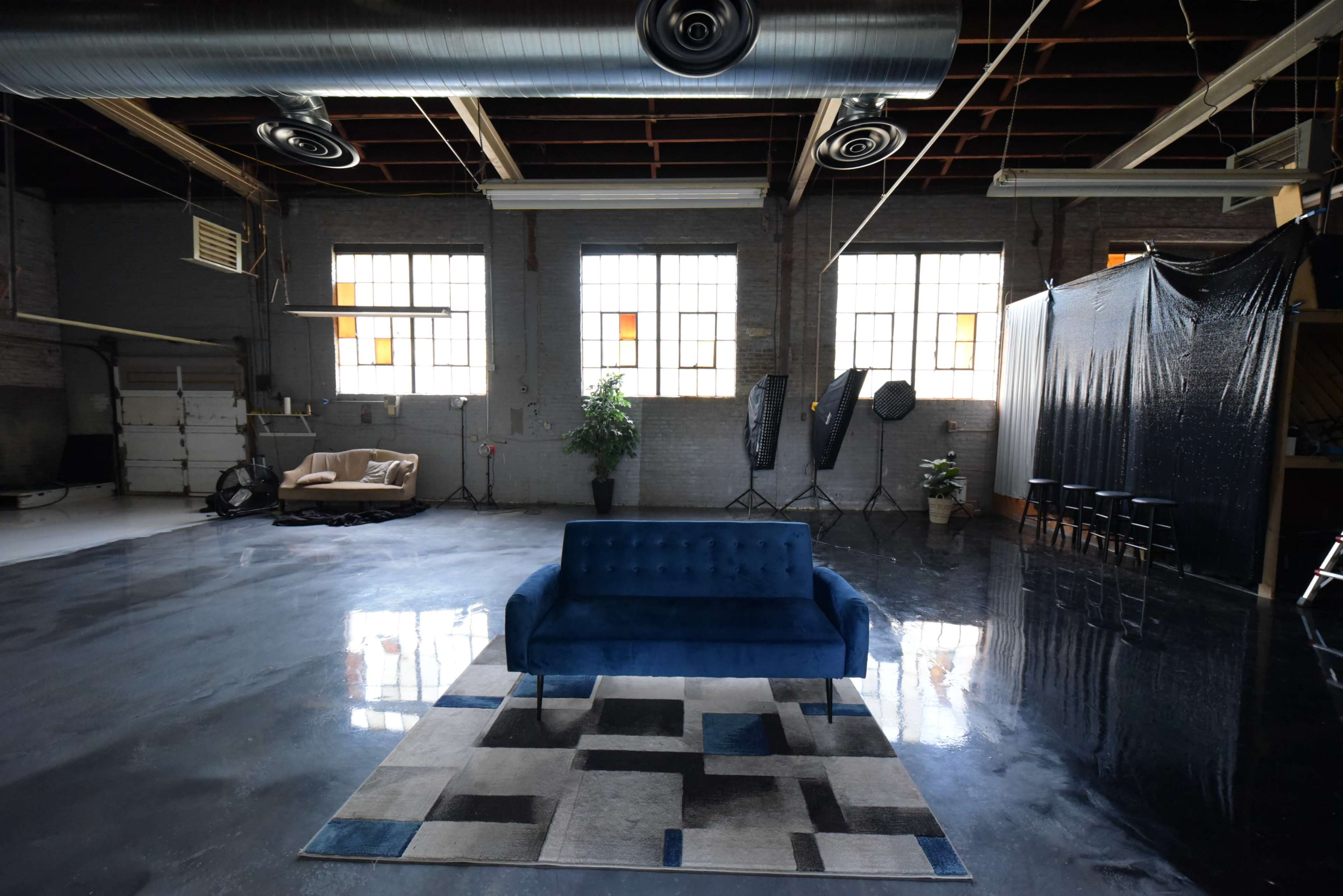 A spacious industrial room with polished concrete floors, large windows, a blue sofa on a patterned rug, and a backdrop set up against one wall.