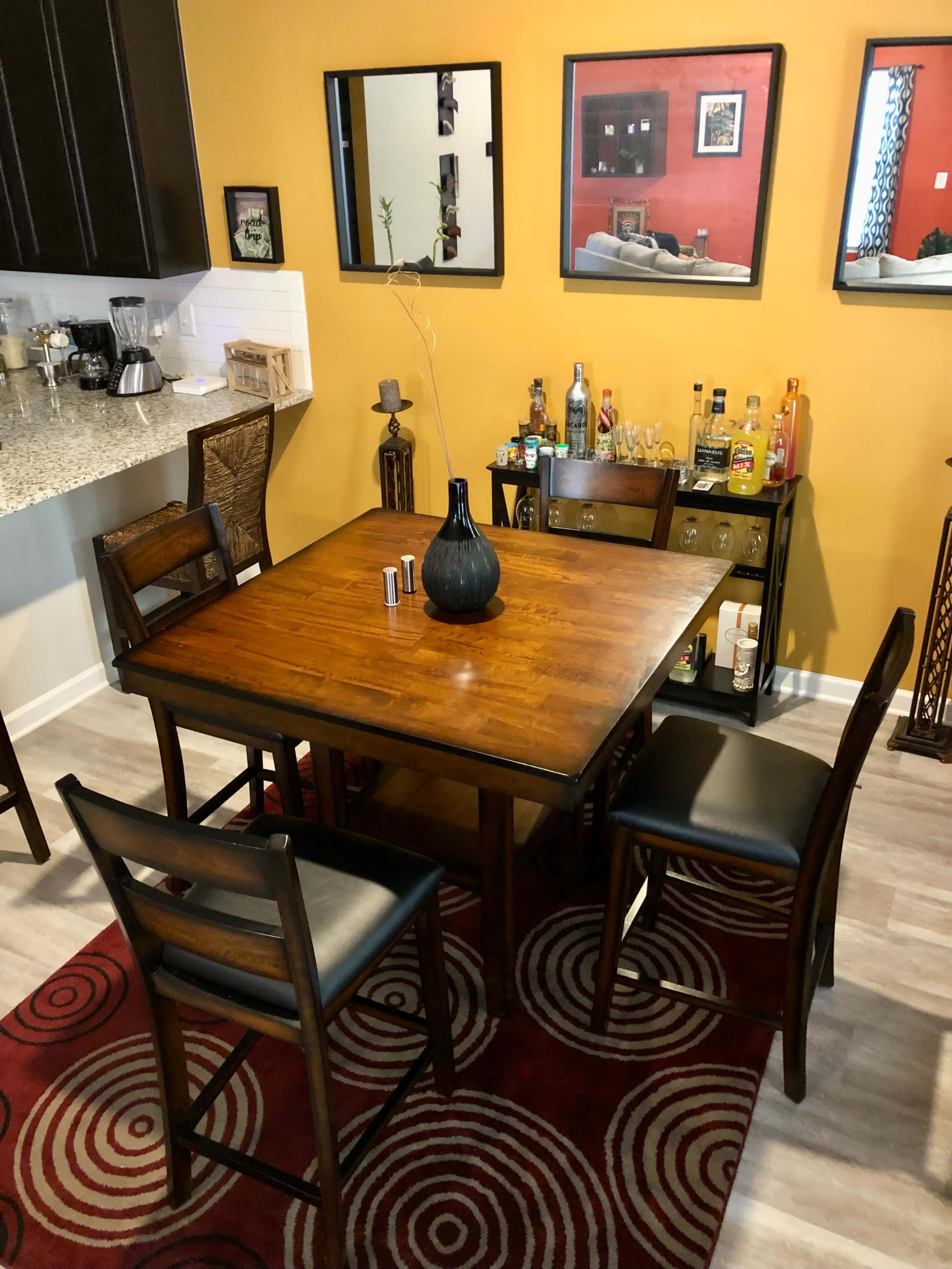 A wooden dining table with four chairs is set in a room featuring yellow walls and several framed mirrors, while a bar cart stocked with various bottles is visible nearby.