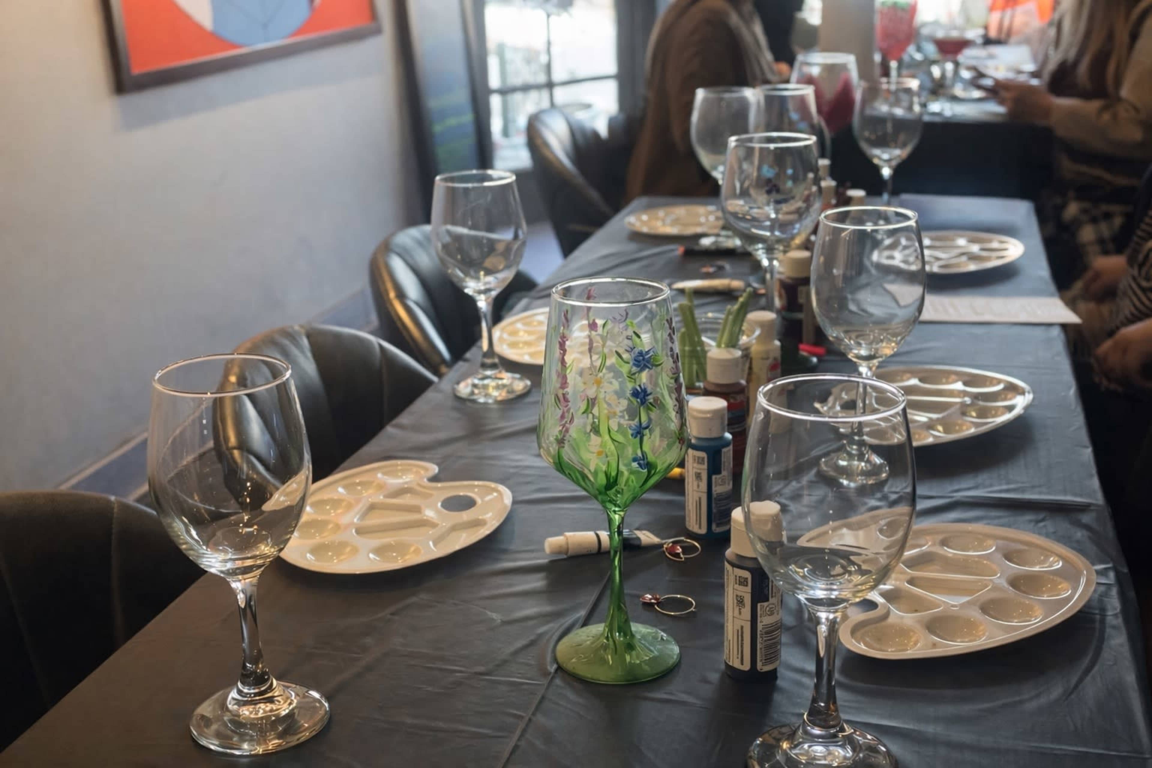 The table is set with empty wine glasses, paint palettes, and art supplies, ready for a gathering or painting event.