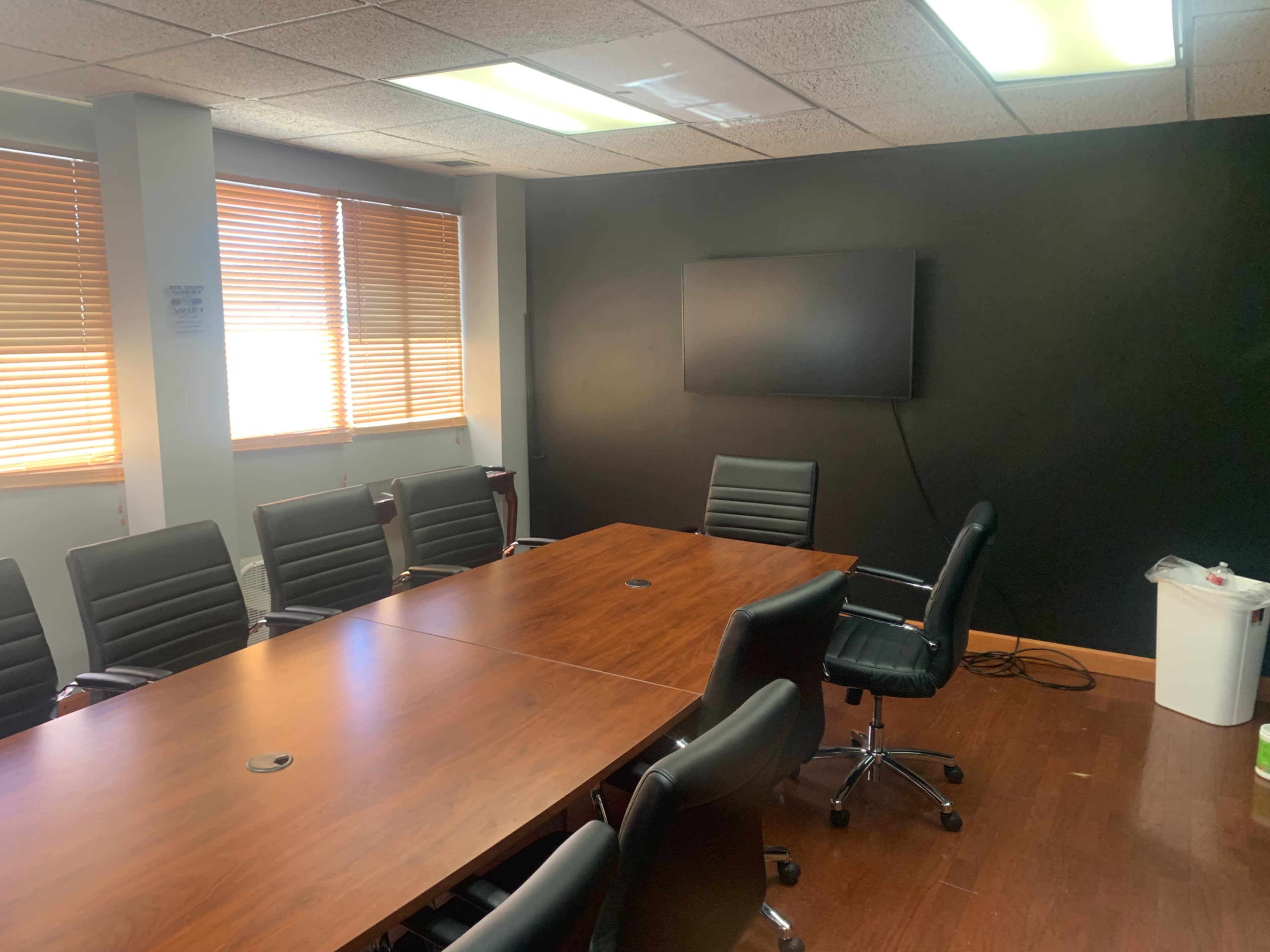 The image shows a modern conference room with a long wooden table, several black leather chairs, a flat-screen TV mounted on a black wall, and large windows with wooden blinds.