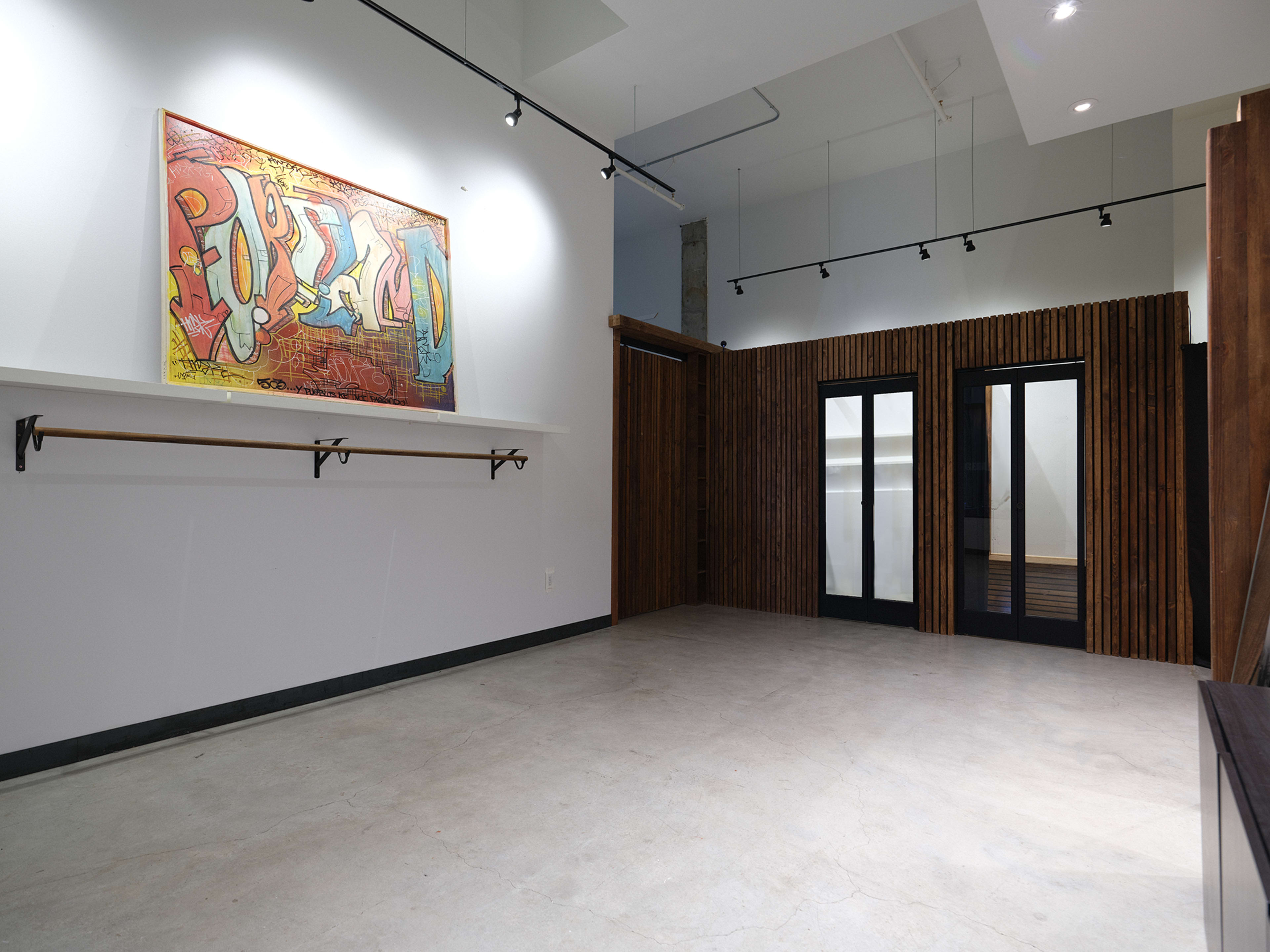 The image shows a spacious, modern room with concrete flooring, a colorful piece of artwork on the wall, and a wooden partition featuring two glass doors.