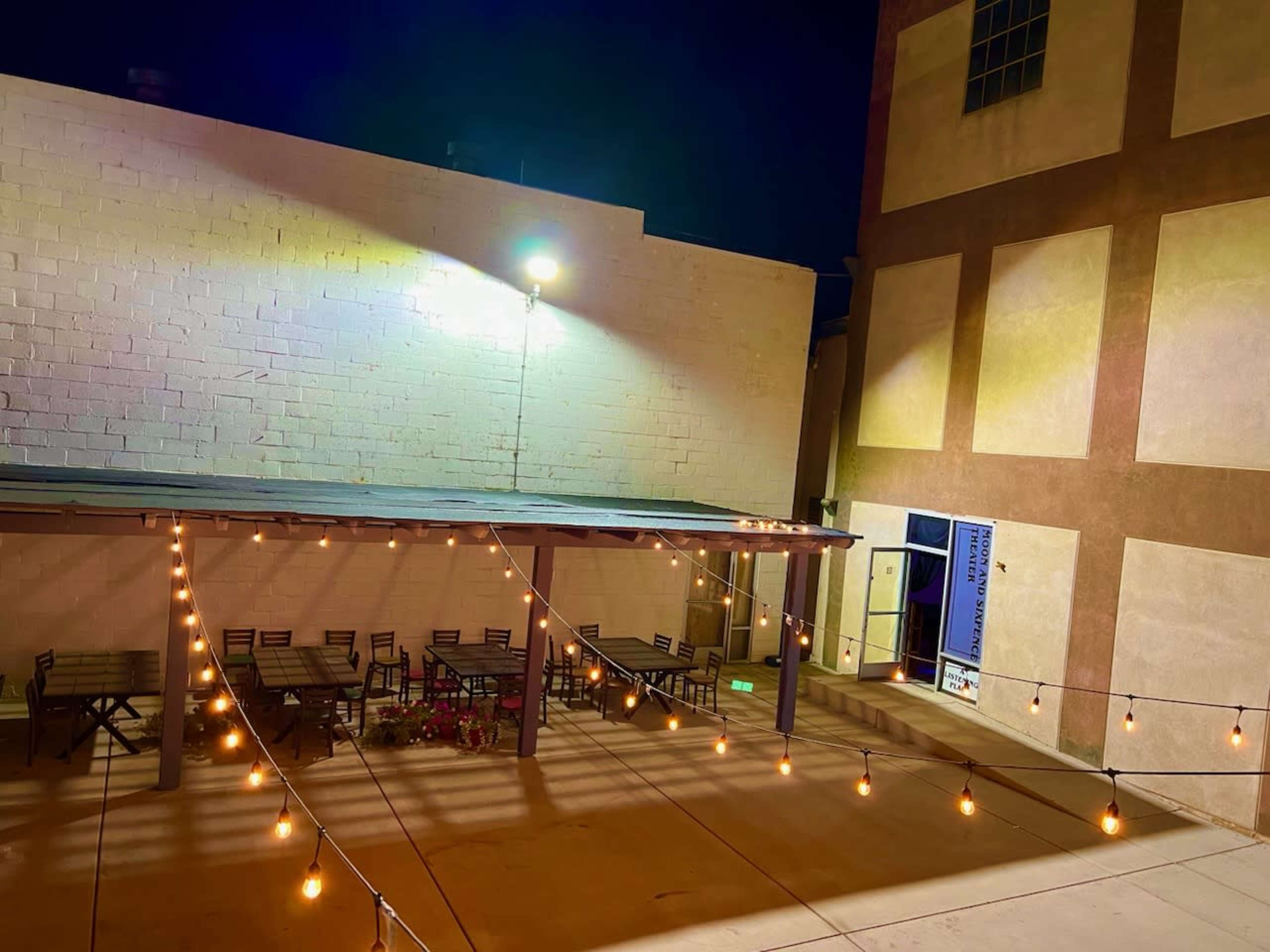 A dimly lit outdoor patio area with string lights and tables under a sheltered roof beside a building.