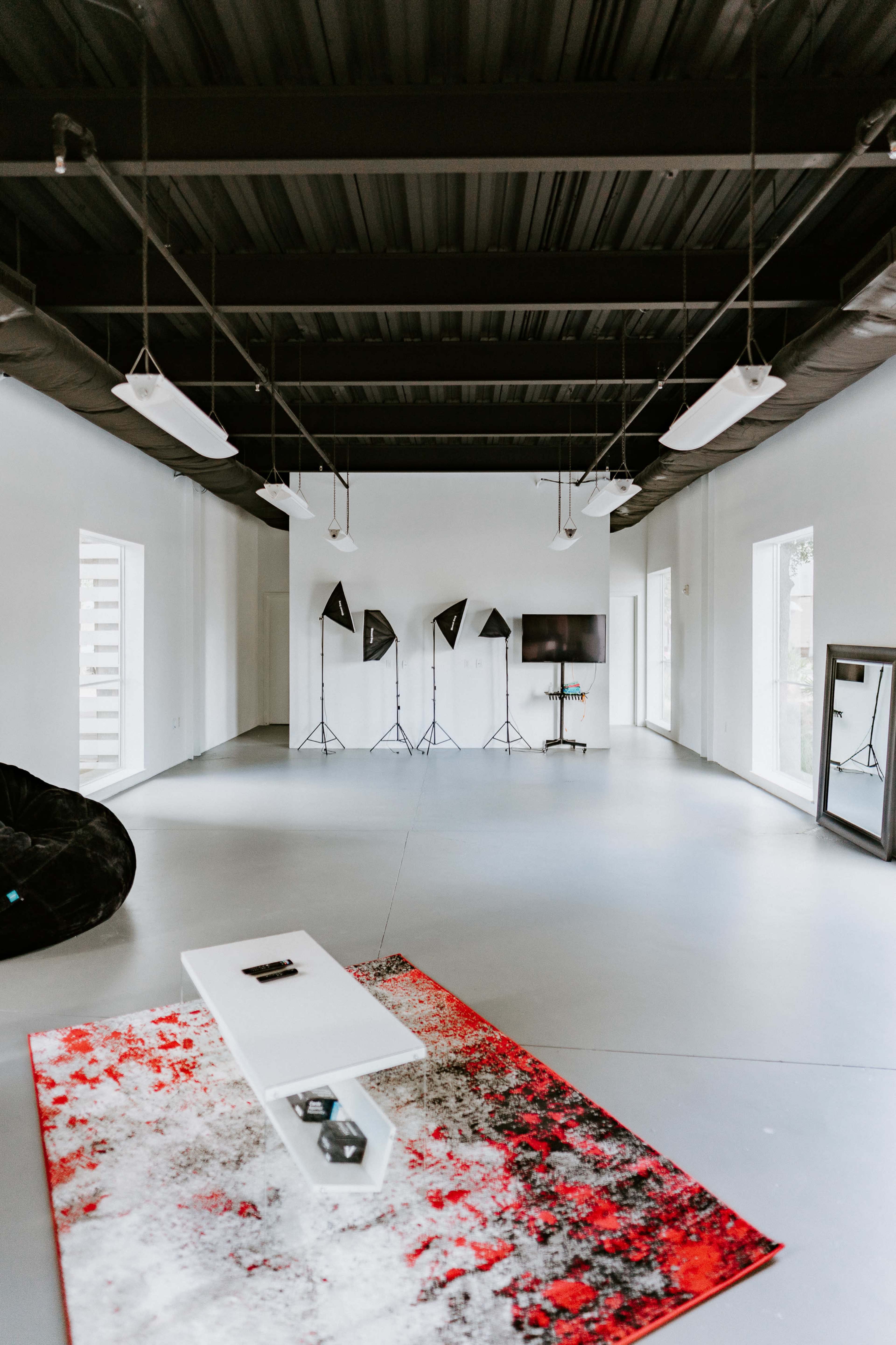 A spacious, minimalist room with gray floors, white walls, and a large red and white rug, featuring several photography studio lights and a screen in the background.