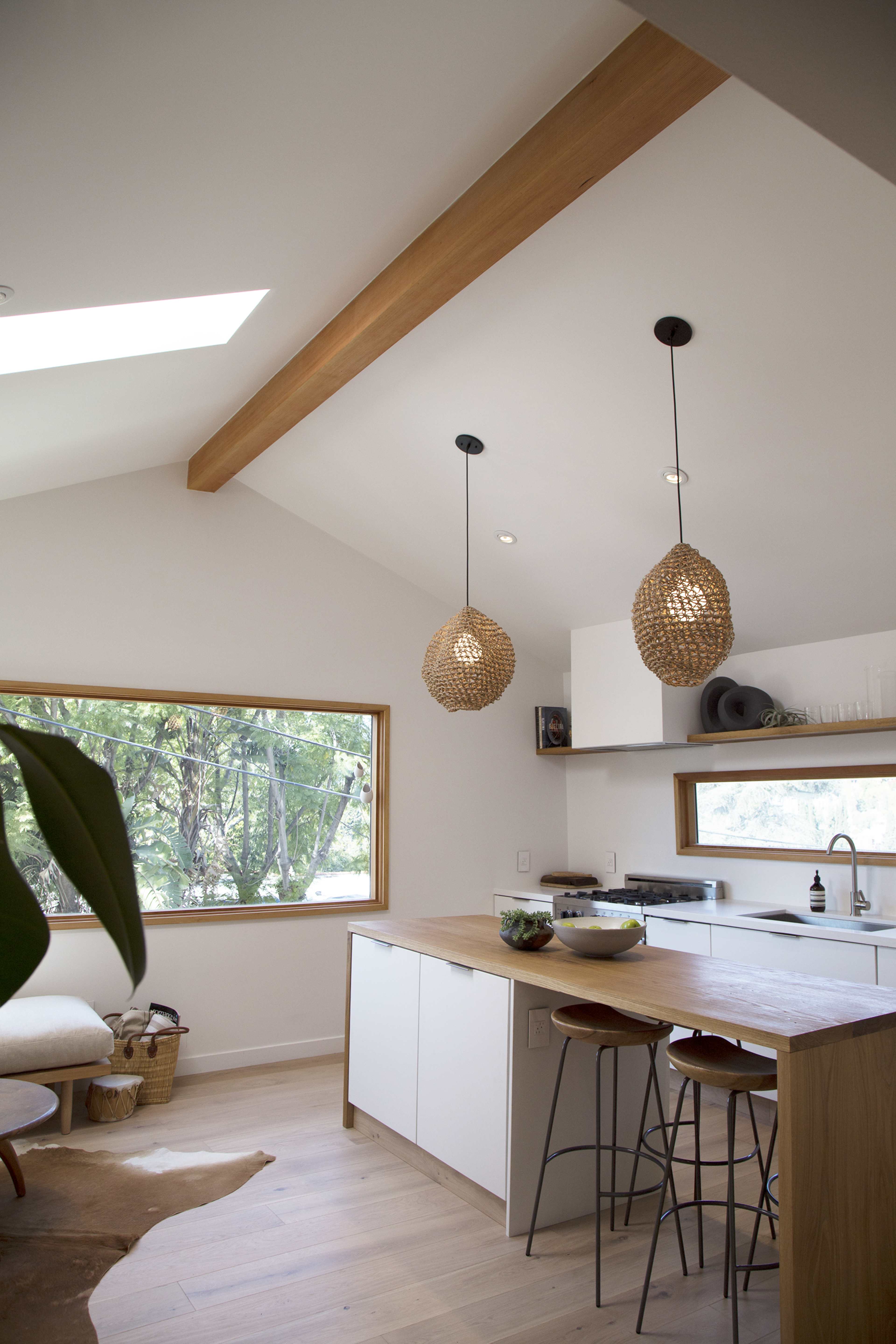 A modern kitchen with a wooden beam ceiling, two woven pendant lights, and a large window overlooking greenery.