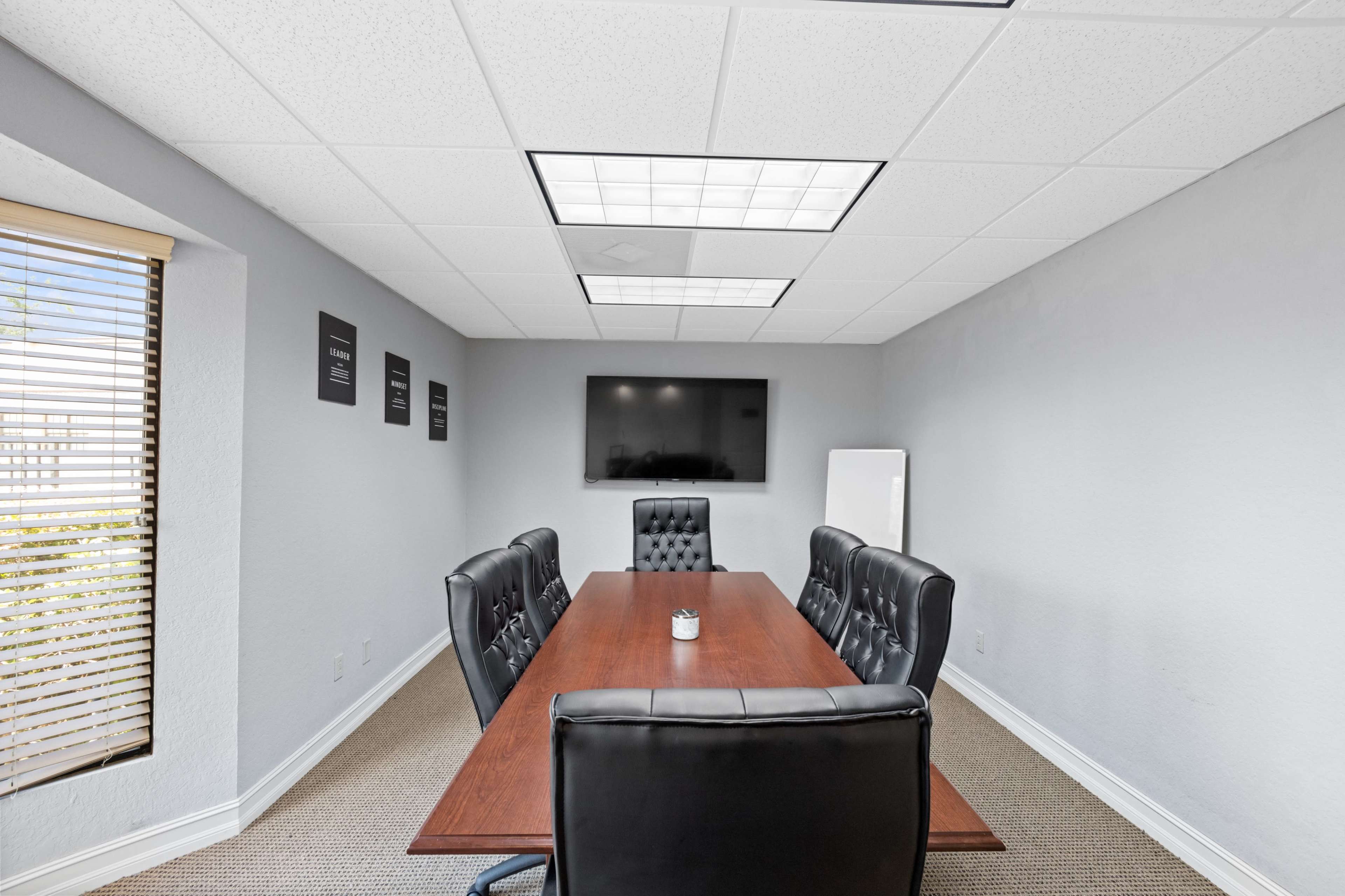 A modern conference room features a long wooden table surrounded by black leather chairs, with a television mounted on the wall and windows letting in natural light.