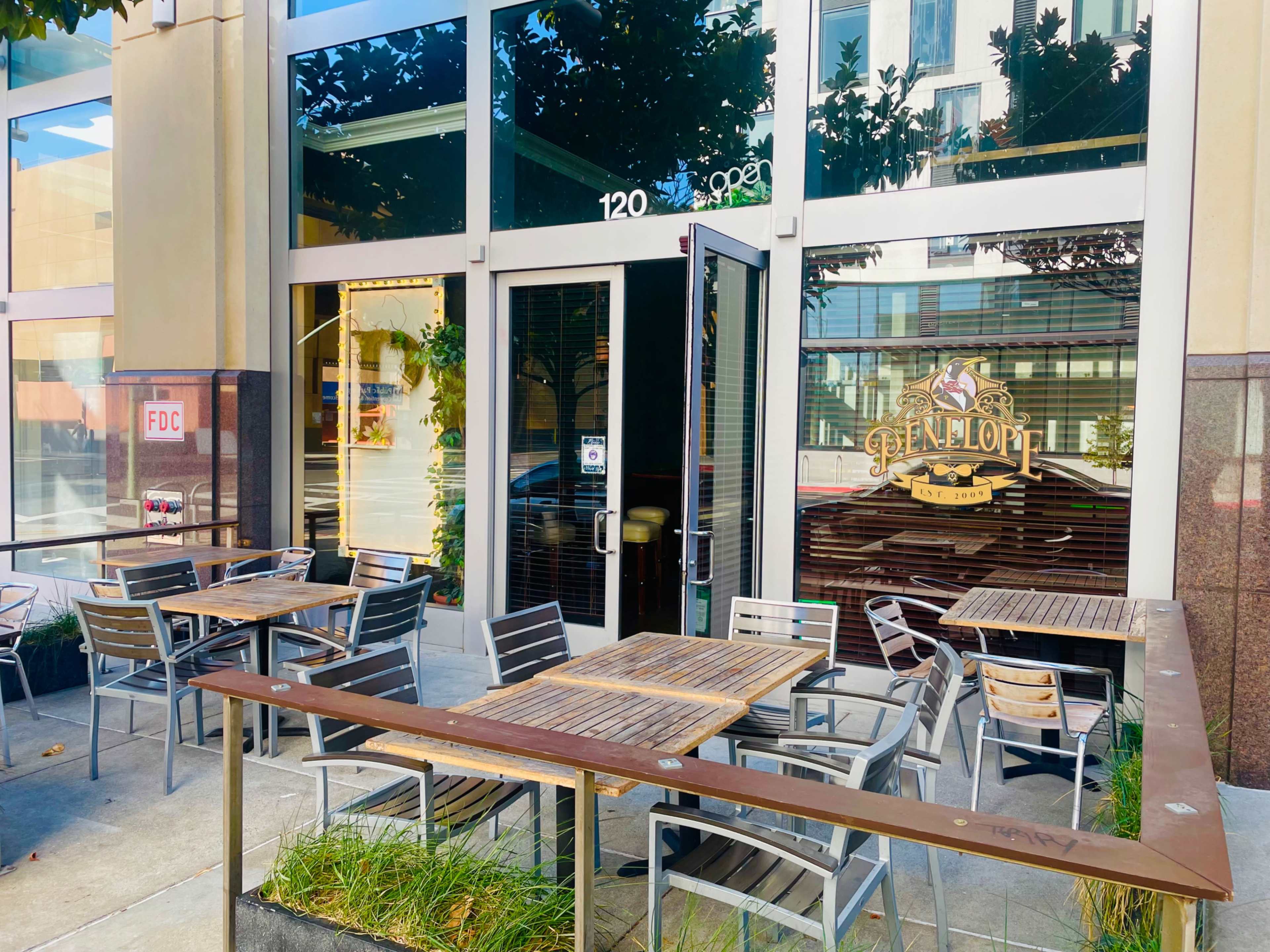 The image shows an outdoor seating area of a restaurant with metal tables and chairs arranged on a sidewalk in front of a glass entrance.