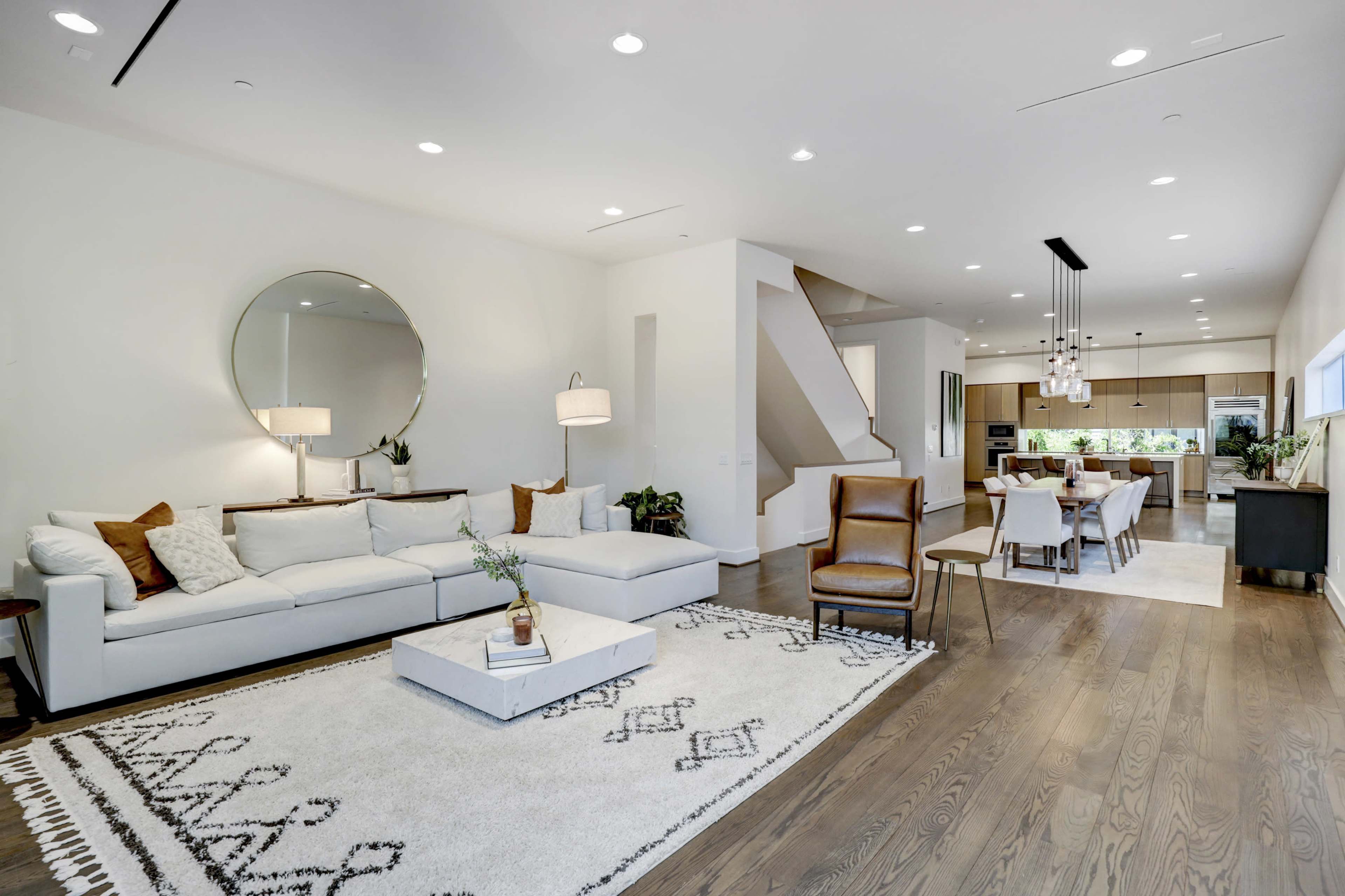 A modern living room with a white sectional sofa, a round mirror, and wooden flooring, adjacent to a dining area and kitchen.