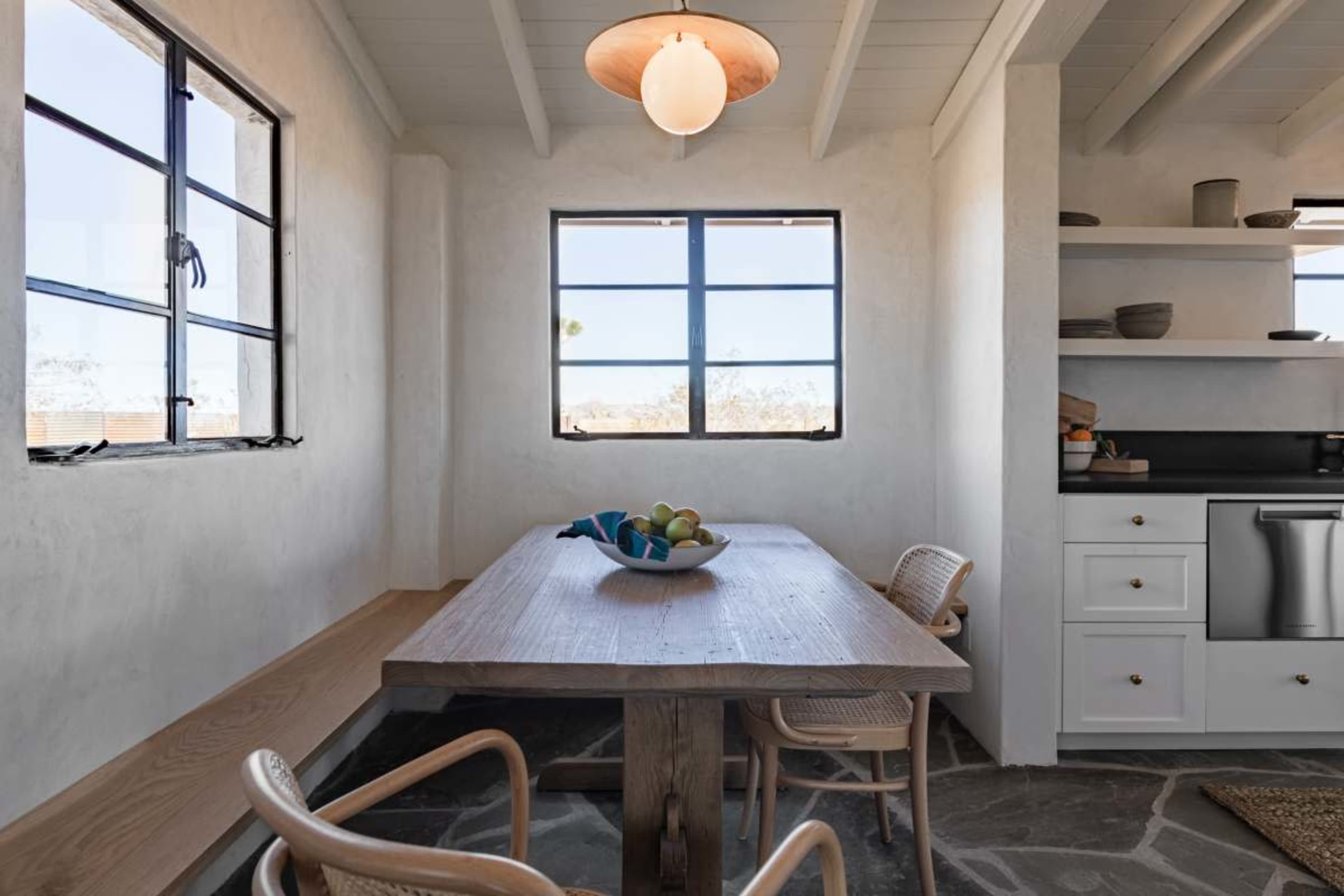 A wooden dining table with fruit sits in a bright kitchen illuminated by overhead lighting, featuring large windows and simple cabinetry.