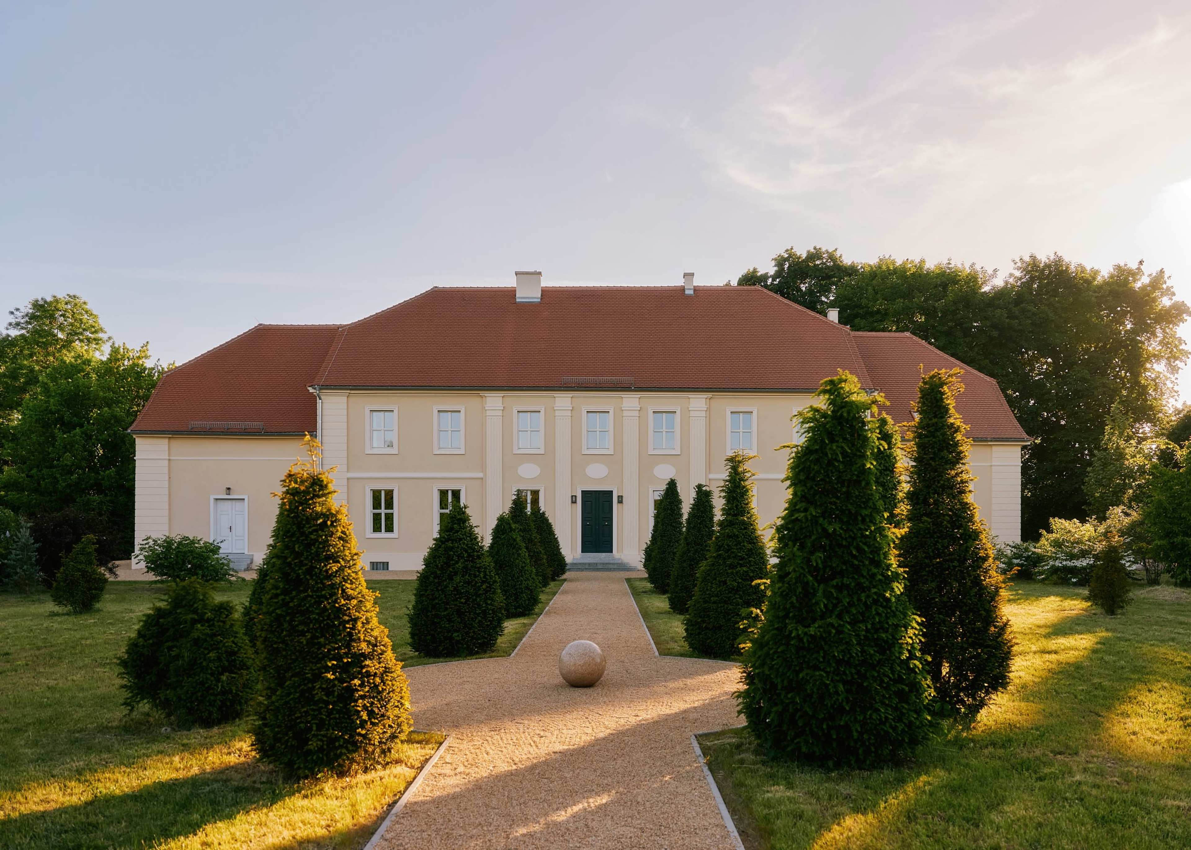 A large, two-story house with a red-tiled roof is surrounded by manicured hedges and a gravel pathway leading to the entrance.