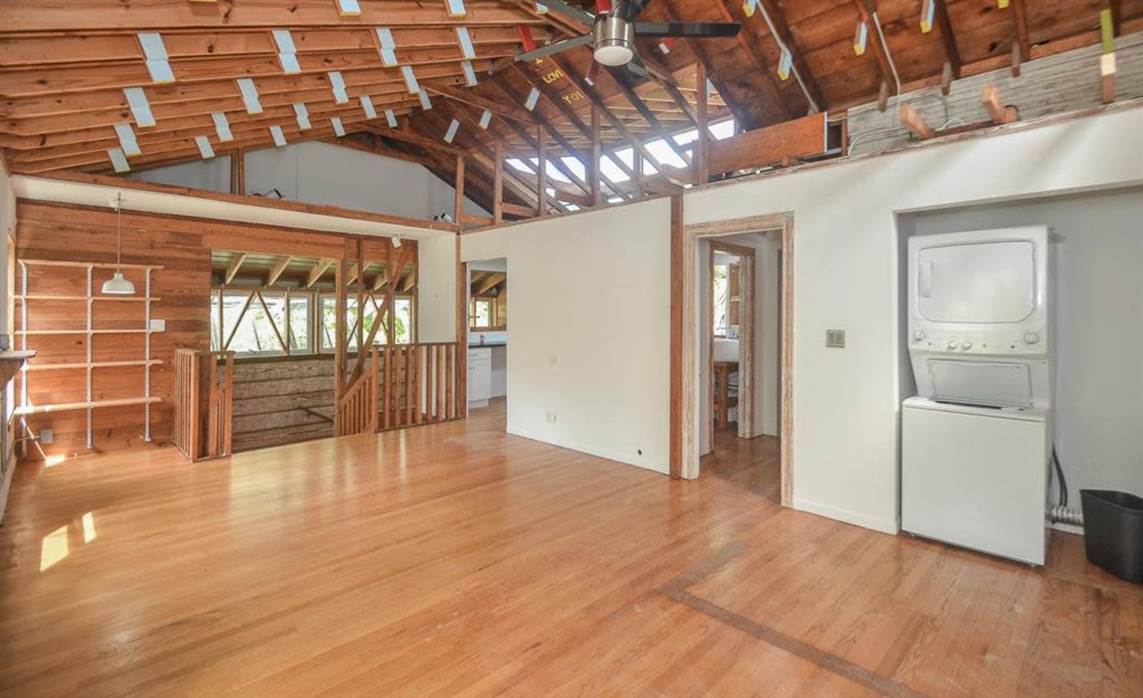The image shows an open-concept living space with exposed beams, wooden flooring, and a washer and dryer set against a white wall.