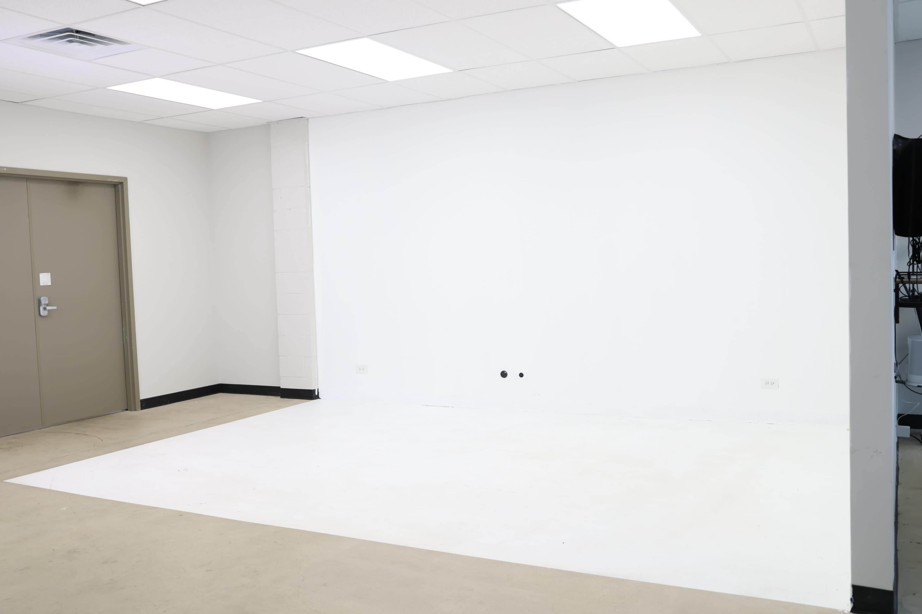 A spacious white room with a light colored floor, a closed door on the left, and an empty wall on the right.